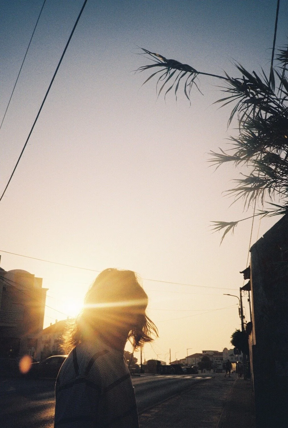 Jelle Rouquart - music producer. Silhouette of a woman with shoulder-length hair standing on a street at sunset, with the sun behind her head, casting a warm glow. There are palm trees and buildings along the street.