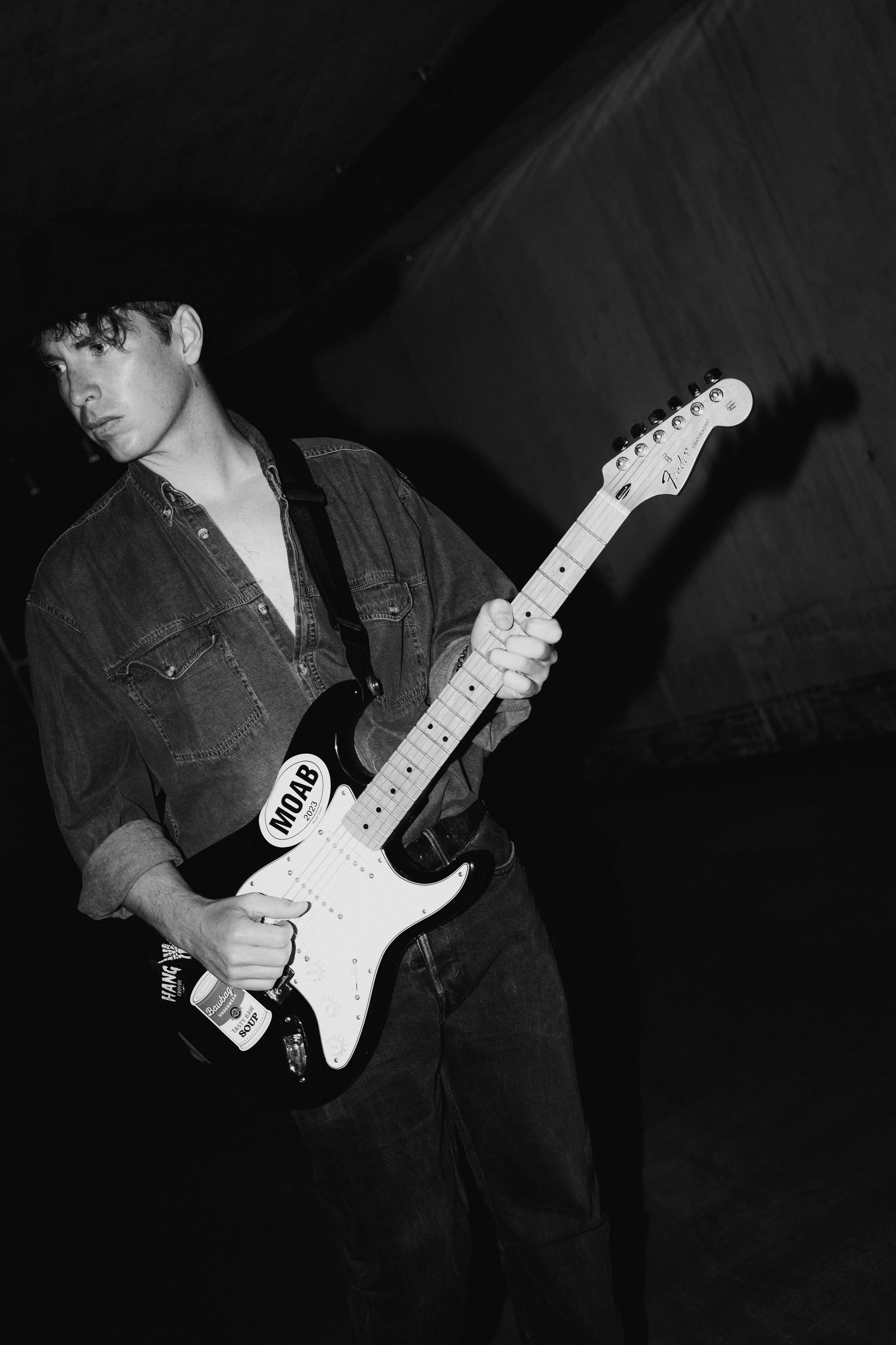A young man wearing a dark cowboy hat, denim jacket, and jeans is holding a Fender Stratocaster electric guitar. The background is dark and plain.