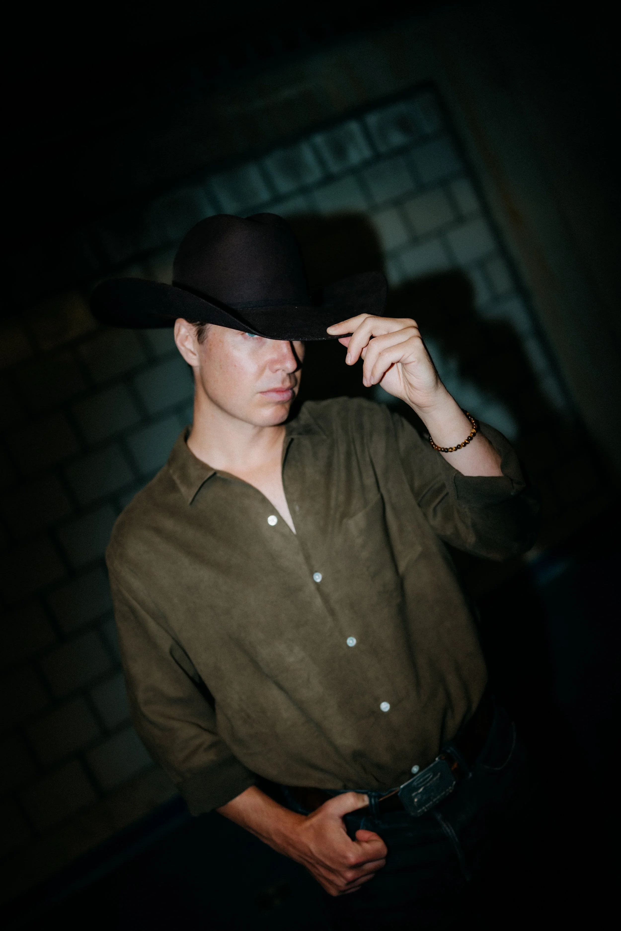 A young man wearing a cowboy hat and a green button-up shirt, standing in front of a brick wall, looking downward and adjusting his hat.