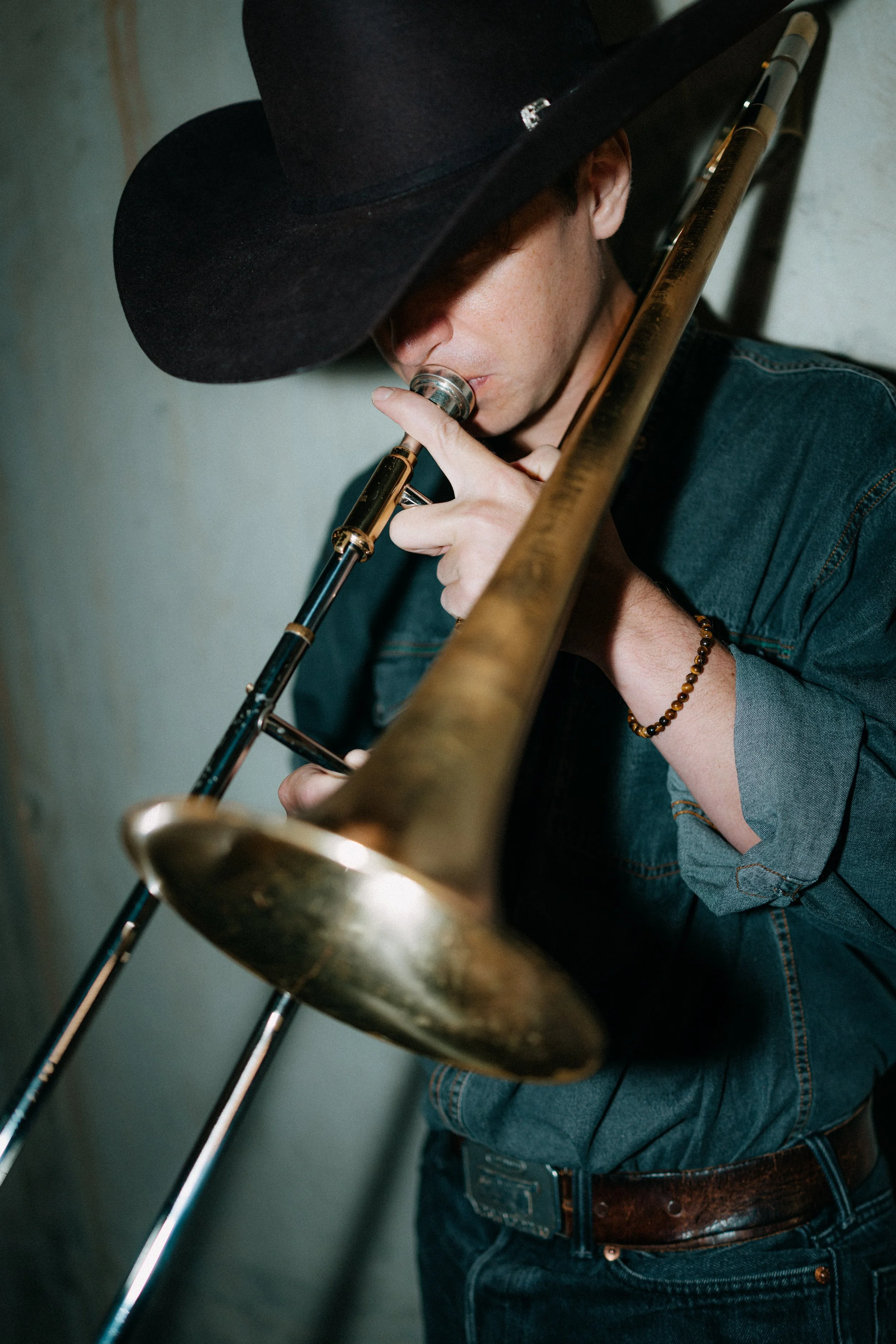 Person wearing a cowboy hat and a denim shirt, playing a brass trombone.