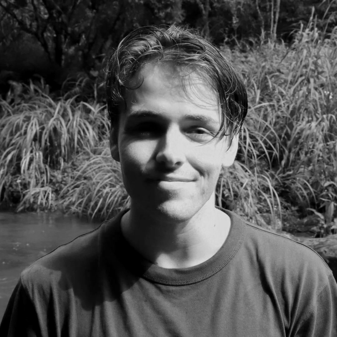 Black and white photo of a young man with wet hair, smiling softly, wearing a T-shirt, outdoors with tall grass and trees in the background.