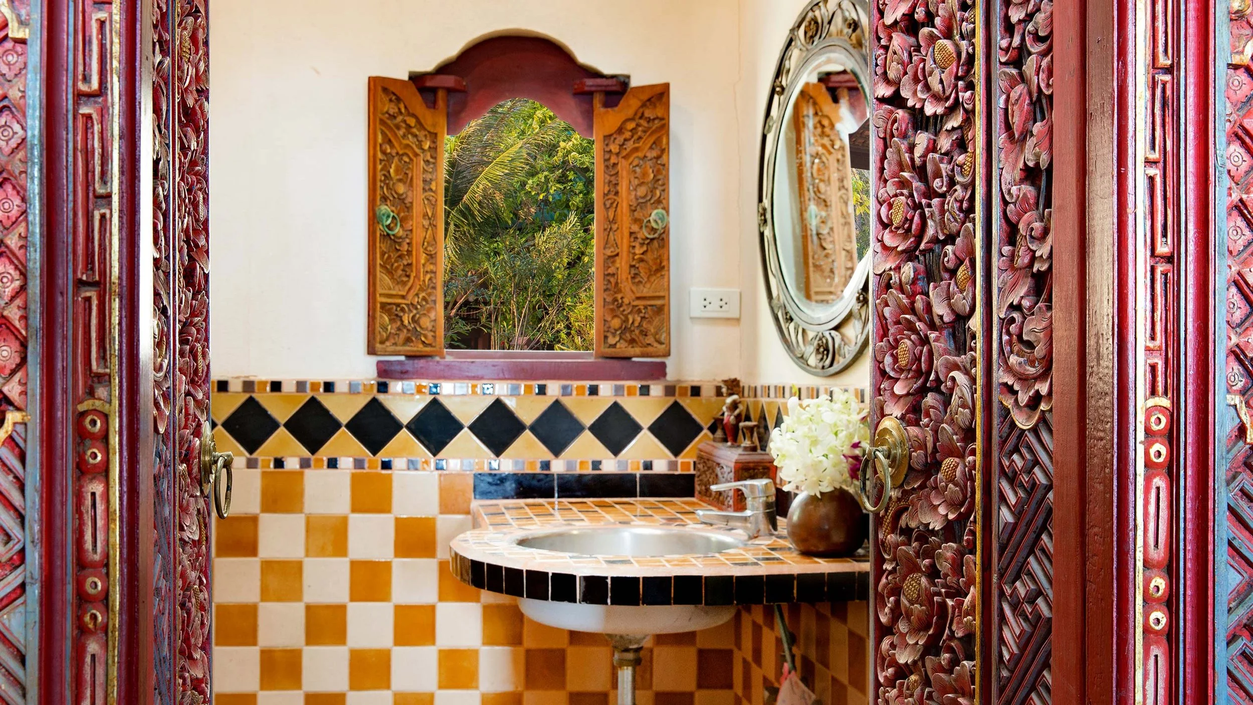 Decorative bathroom with carved wooden doors, patterned tiles, a small sink, a mirror, and an open window showing greenery outside.