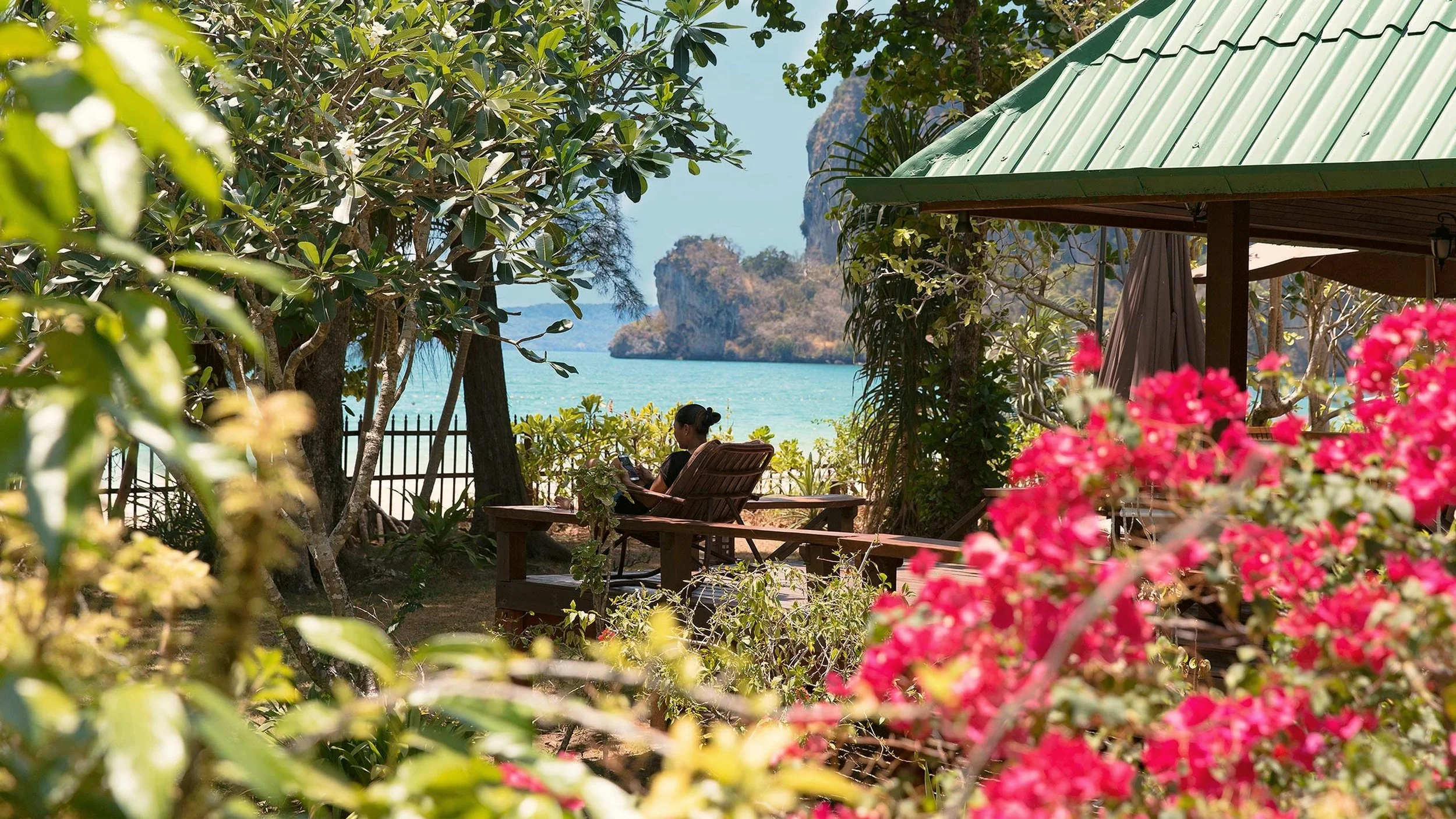 A person sitting on a wooden lounge chair on a tropical beach, surrounded by greenery and pink flowers, with turquoise water and rocky islands in the background at Railay Beach, Thailand
