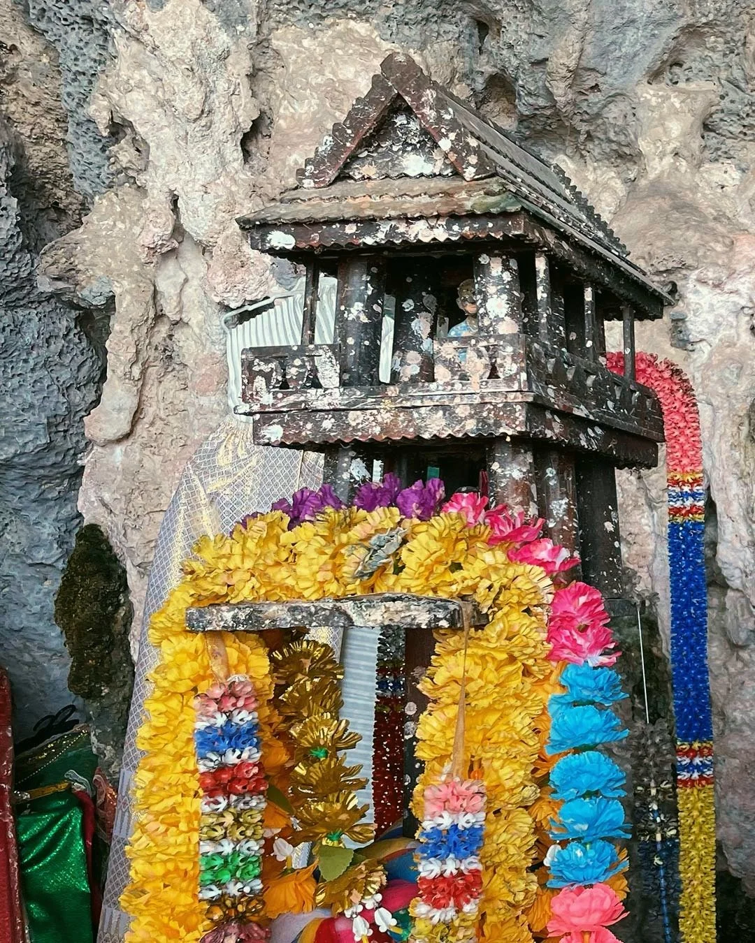 Small wooden shrine or house surrounded by colorful flower garlands and decorations, set against a rugged stone wall.