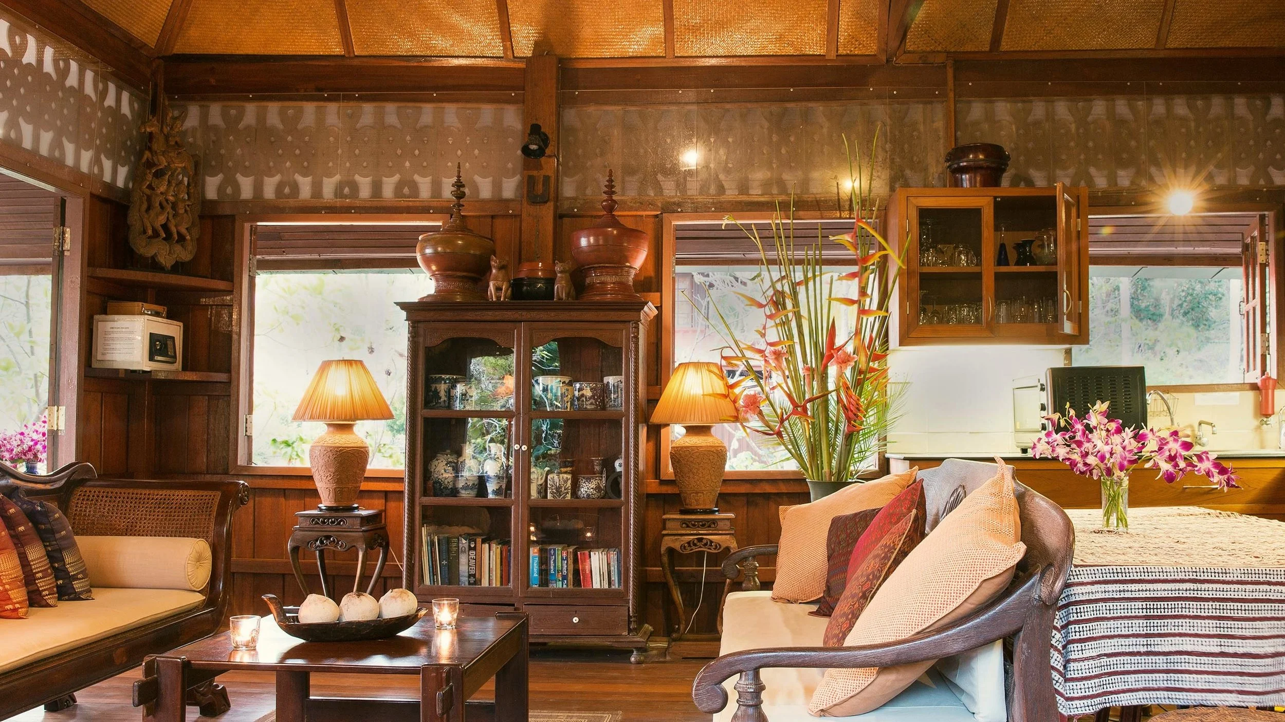 A cozy living room with wooden walls and furniture, decorated with lamps, flowers, and books, with large windows letting in natural light.