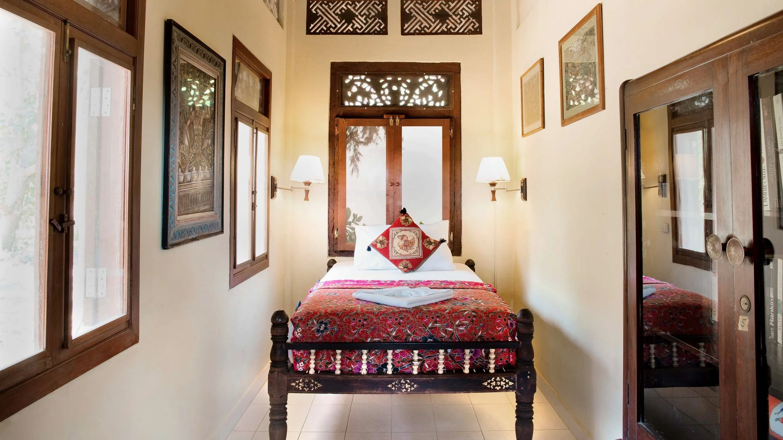 A cozy bedroom with a single bed featuring a patterned red bedspread and a decorative pillow, positioned against a wall with windows on both sides, wooden frames, and artwork, illuminated by wall-mounted lamps.