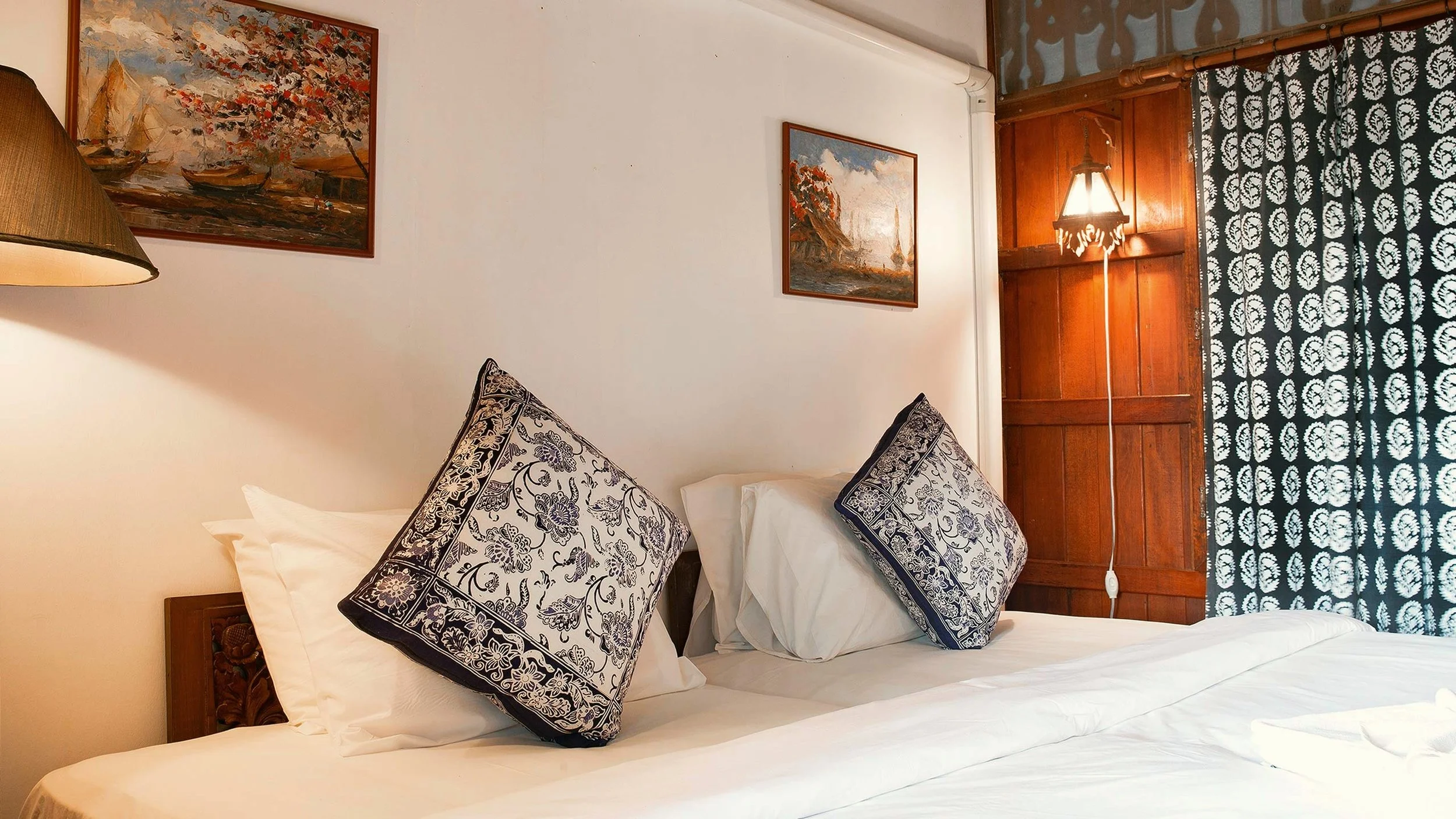 A cozy hotel bedroom with two decorative pillows featuring intricate black and white floral patterns, a wooden headboard, a nightstand with a lamp, painted landscape artwork on the walls, wooden paneling, a lamp affixed to the wall, and patterned curtains.