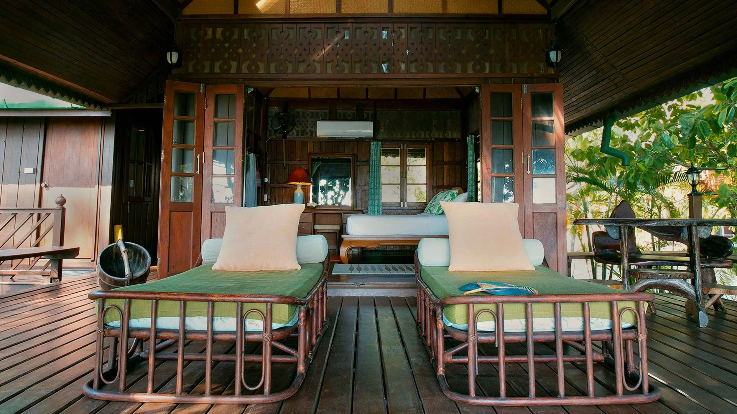A cozy wooden cabin interior with two bamboo lounge chairs with green cushions and beige pillows on a wooden deck, overlooking a bedroom area with a single bed, bedside lamp, small table, and windows with green curtains, surrounded by lush trees.