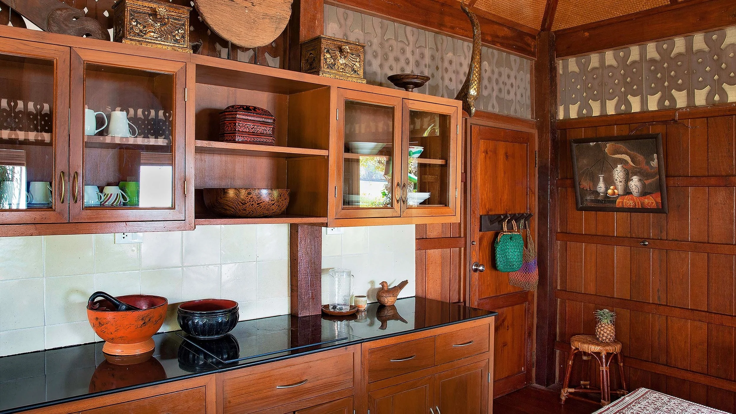 Kitchen with wooden cabinets, glass-fronted upper cabinets, and ceramic bowls and cups on the counter. Decorative items and artwork are on the walls and shelves, including a painting of vases and a carved pineapple sculpture.