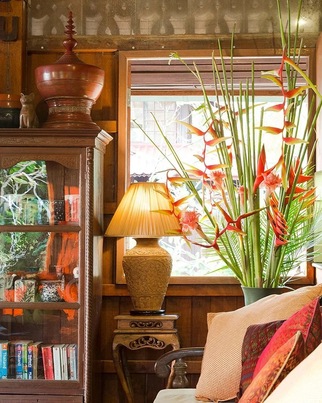 Interior of a cozy room with a large potted flower arrangement of pink and orange flowers, a warm table lamp on a carved wooden side table, a glass-fronted wooden cabinet filled with books and decorative items, a window letting in natural light, and wooden walls and furniture.