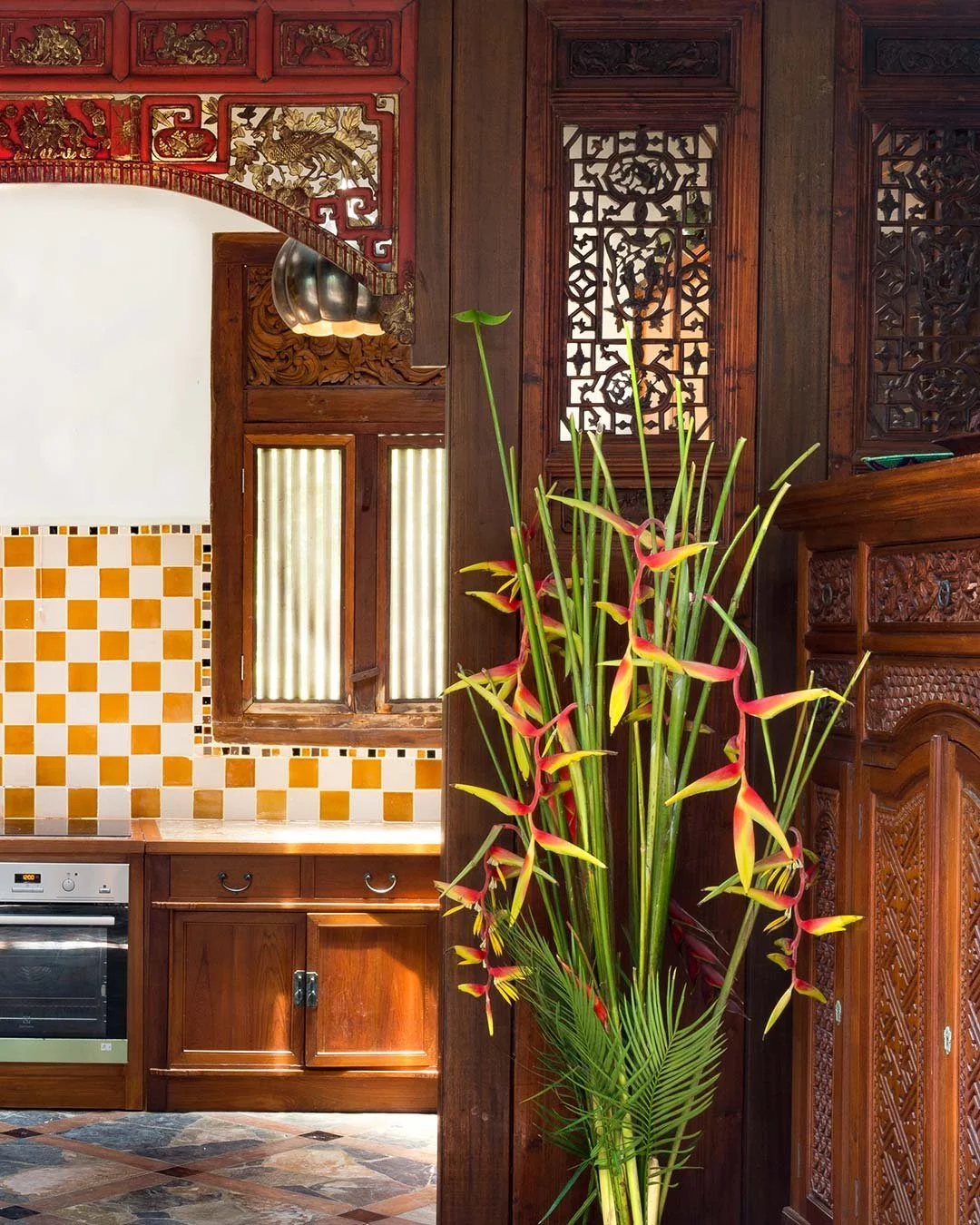 A wooden interior with intricate carvings, a window with frosted glass, a kitchen counter with an oven, and a vibrant arrangement of tall green and red flowers.