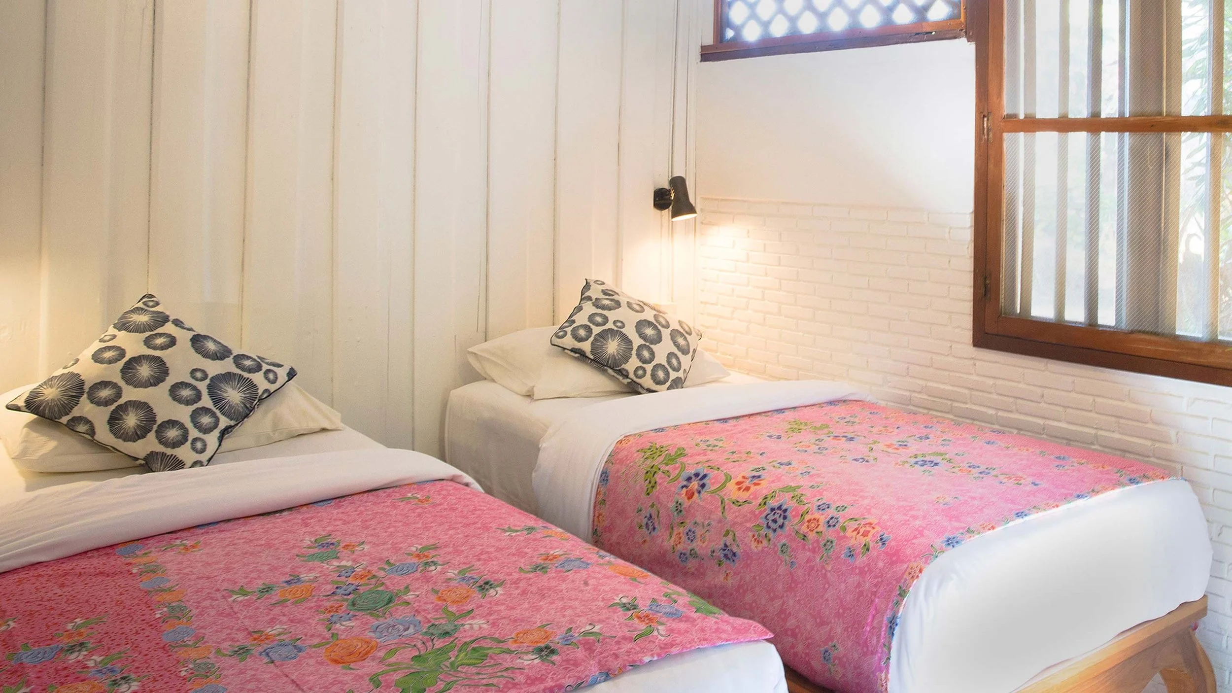 Two beds with floral pink bedspreads and black and white patterned pillows in a bright room with wooden windows and white brick walls.