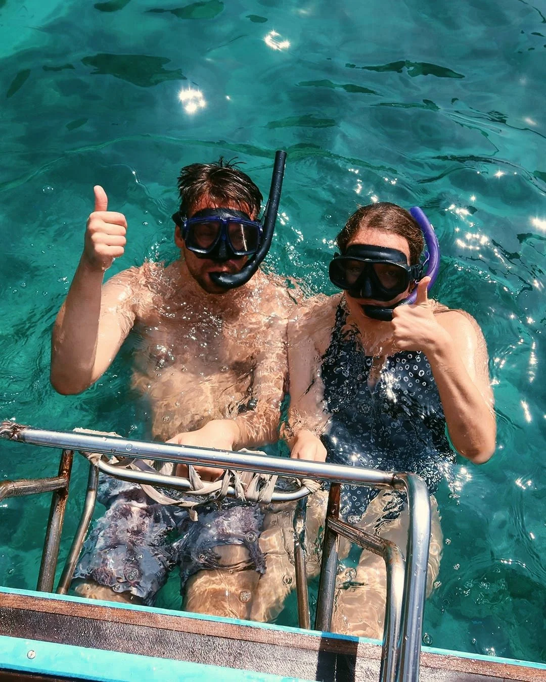 Two people in snorkeling gear giving a thumbs up in clear blue water, standing on a boat ladder at Railay Beach, Thailand
