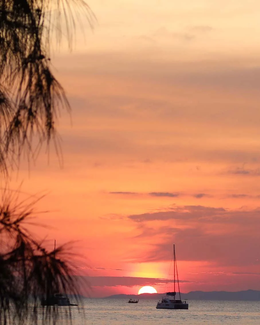 A sunset over the ocean with sailboats and a mountain in the distance, framed by tree branches on the left at Railay Beach, Thailand
