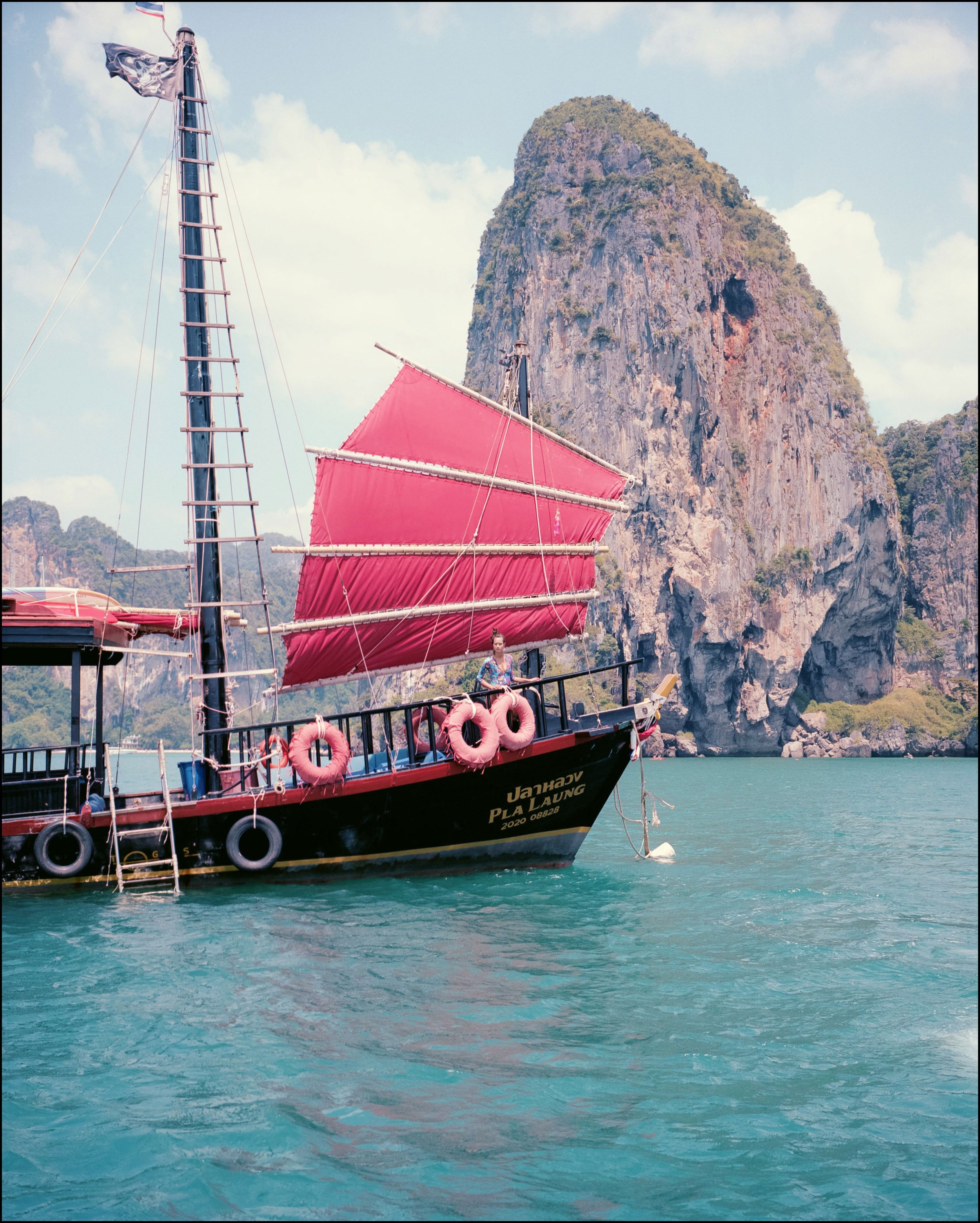 A sailboat with pink sails and black hull, anchored in turquoise water with rocky limestone cliffs in the background.