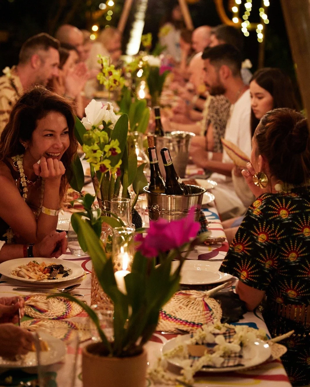 People at an outdoor dinner party seated around a long table decorated with flowers, candles, and plates of food, enjoying conversation and laughter.