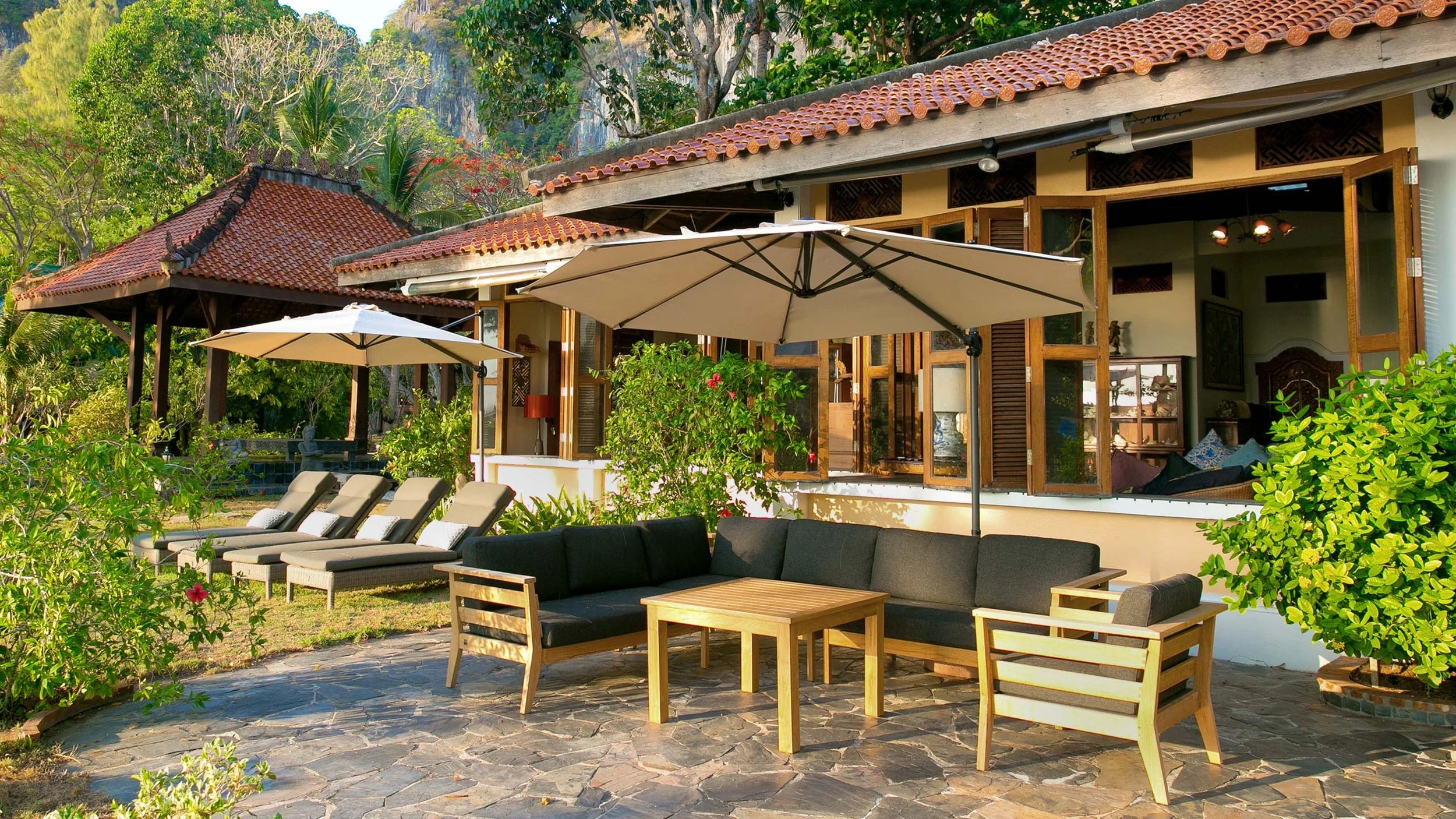 Outdoor patio area with black cushioned sofa and matching armchair, wooden coffee table, two large umbrellas providing shade, and four lounge chairs, surrounded by lush greenery and trees, with open windows of a house in the background.