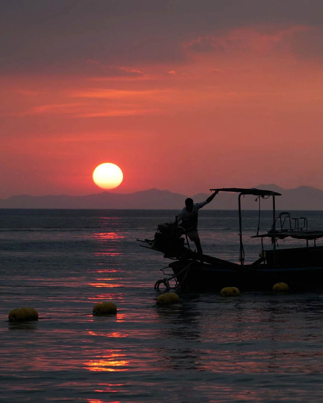 A person standing on a boat during a colorful sunset over the ocean, with distant mountains in the background.