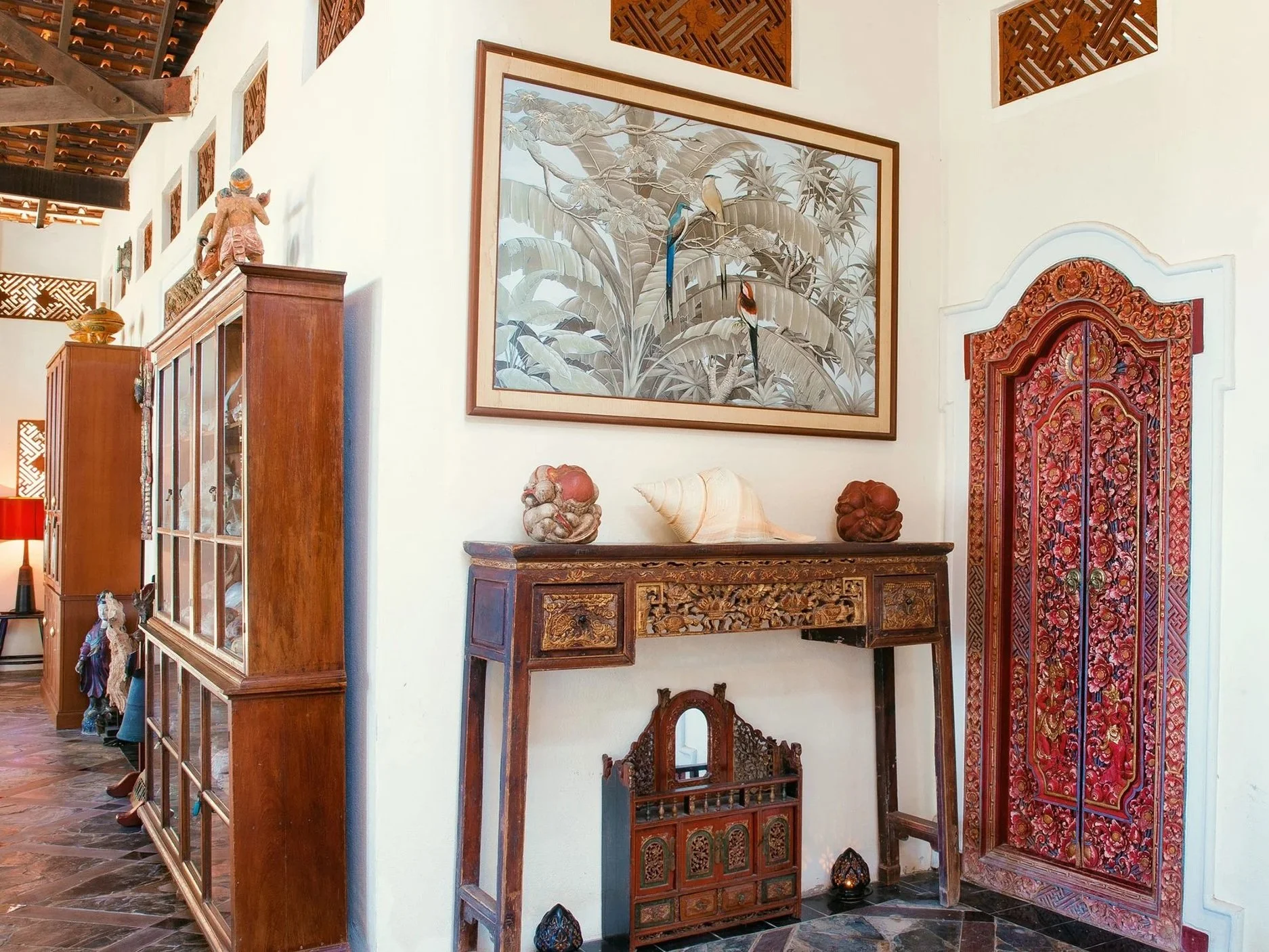 Interior of a room with Asian-style decor, including a carved wooden console table, decorative shells, and a large framed artwork of tropical plants and birds on the wall. A red ornate door with intricate patterns is also visible.