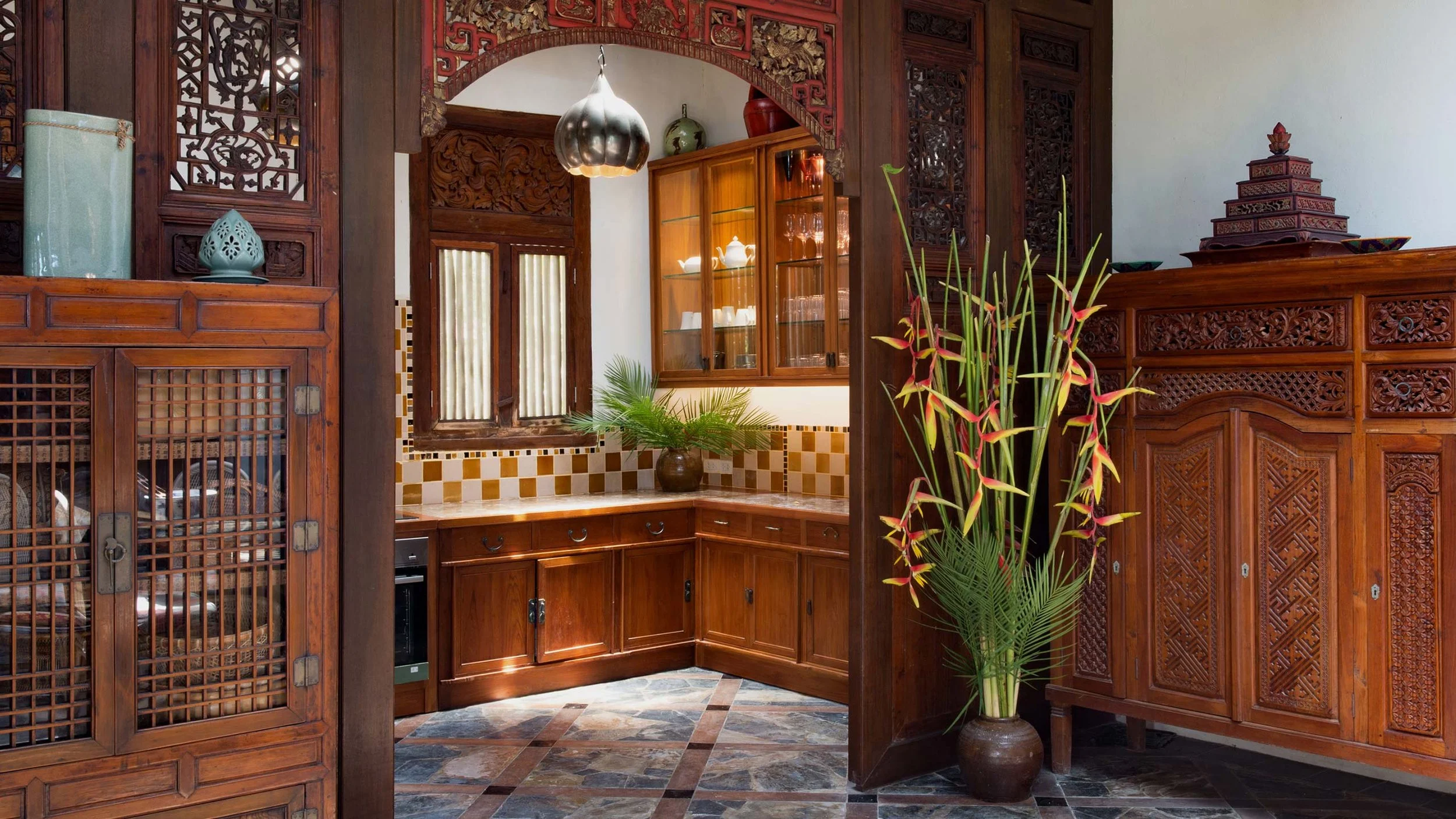 View of a traditionally decorated kitchen with wooden cabinets, decorative wood carvings, and a large vase with tropical plants.