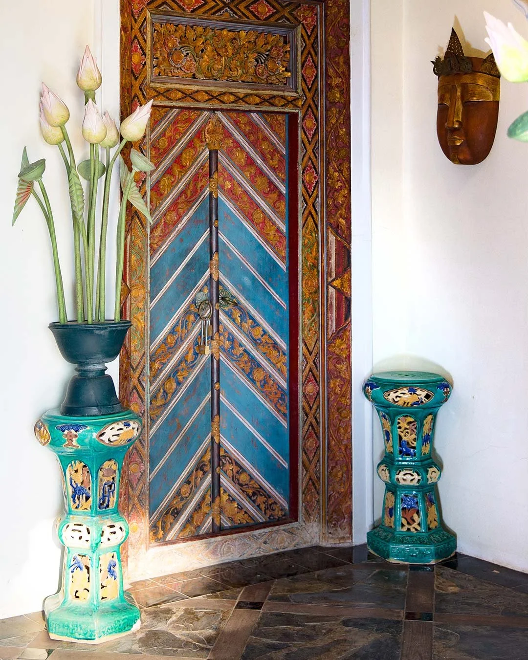 Decorative interior with a colorful carved wooden door, two ornate ceramic vases with floral designs, a tall vase with artificial white lotus flowers, and a wall-mounted wooden mask.