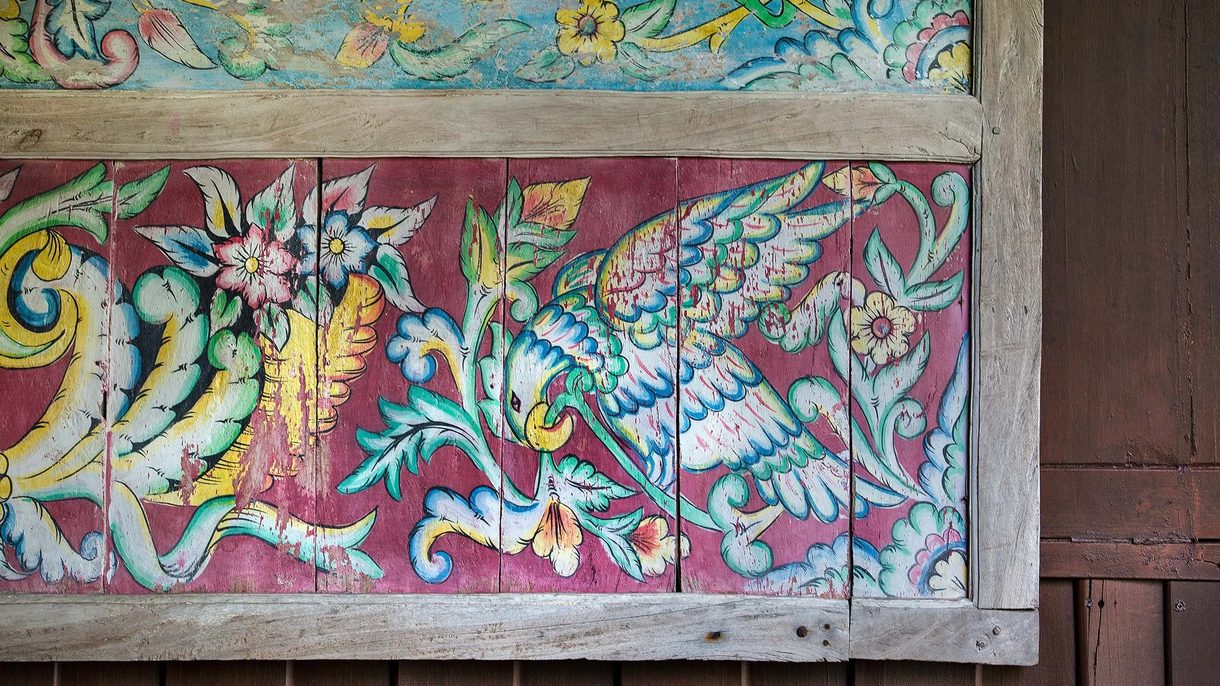 Colorful hand-painted mural featuring a bird with wings spread, surrounded by flowers and decorative swirls, on reddish wood panels framed with weathered wood.