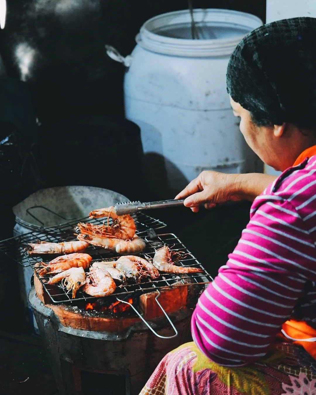 A woman wearing a pink striped shirt and a black hat is grilling shrimp on a small charcoal stove outdoors.