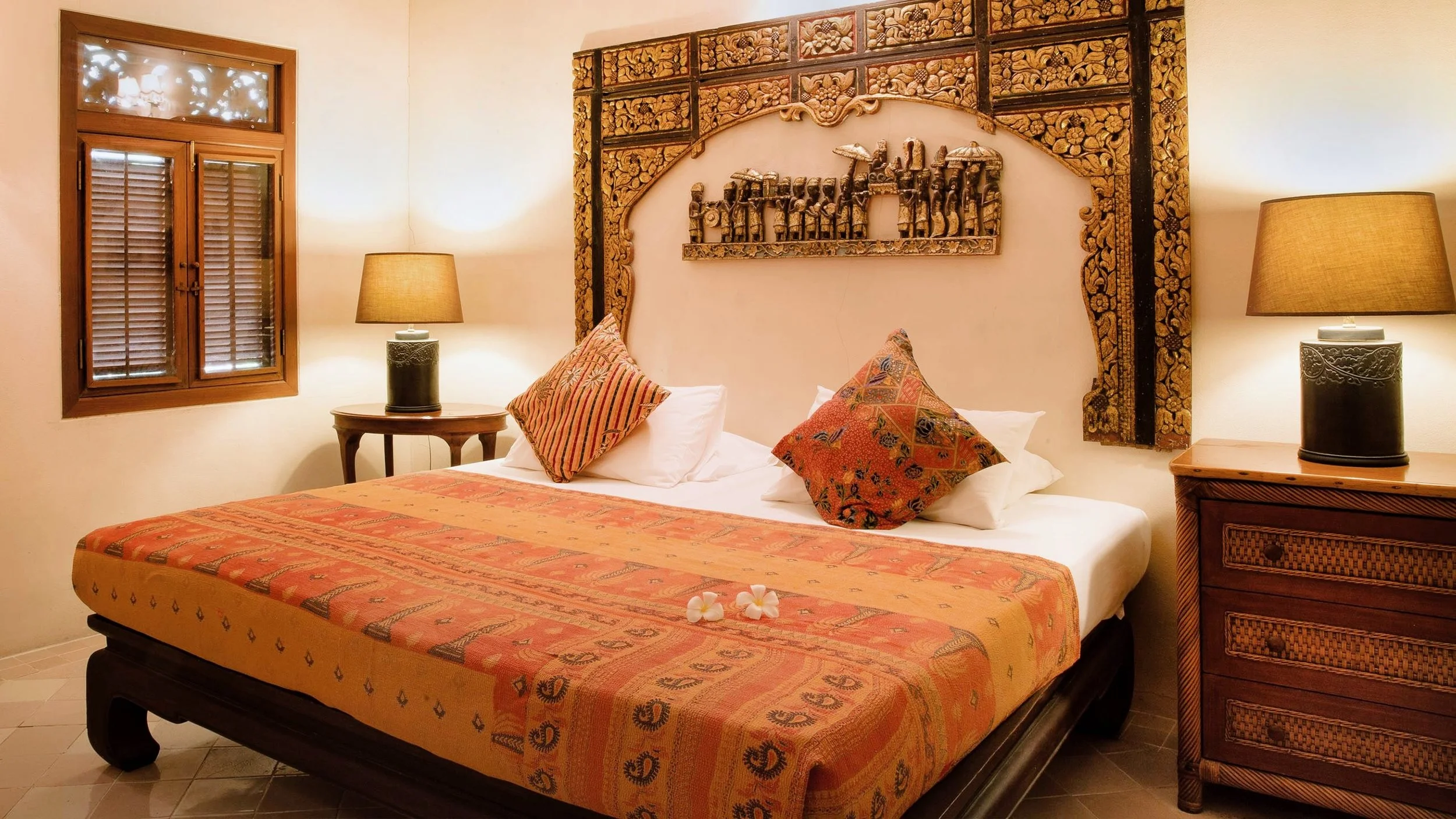 A cozy bedroom with a wooden bed frame and an orange patterned bedspread. There are two decorative pillows on the bed, one striped and one with a floral pattern. A pair of flowers is placed on the bedspread. On each side of the bed, there are wooden nightstands with matching lamps. The wall behind the bed has ornate wooden carvings and artwork. There is a window with wooden shutters and a small window above it with decorative lights.