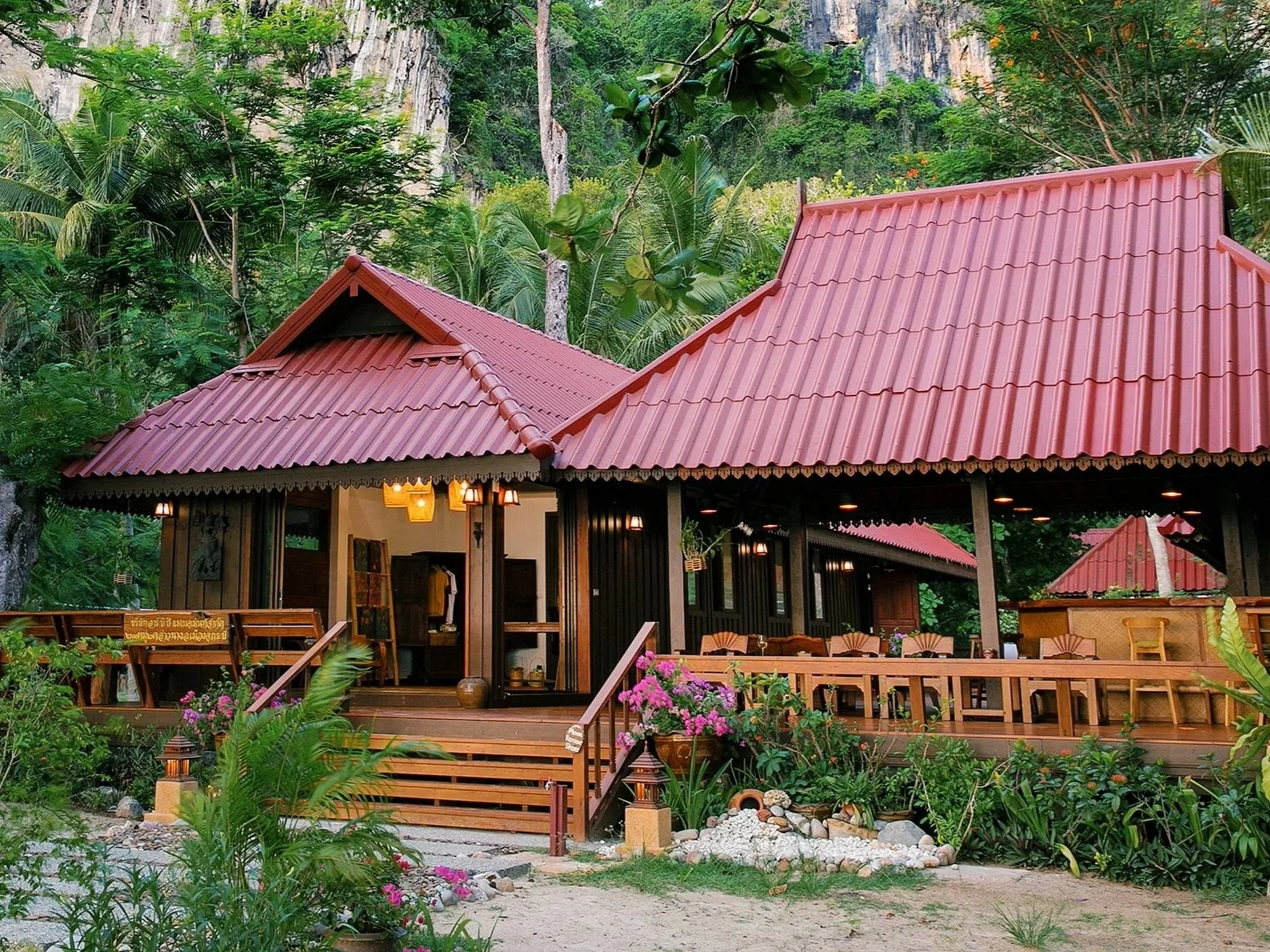 A Thai-style wooden house with a red tile roof, surrounded by lush green tropical plants and trees, with a scenic mountain backdrop.