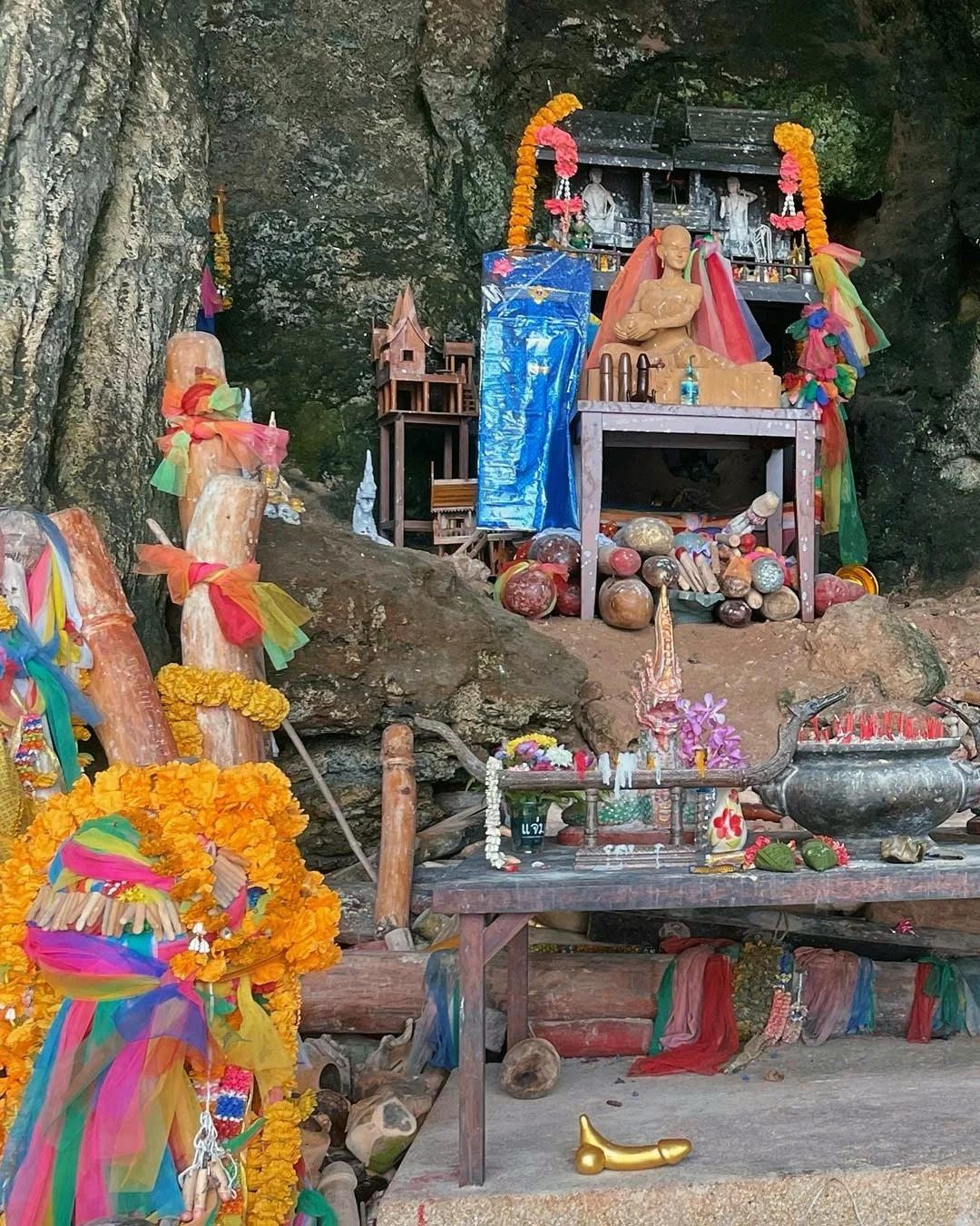 Small spirit shrine or altar set against a cave or rocky wall, decorated with colorful ribbons, flowers, and statues of spiritual figures, with offerings of food and other items.