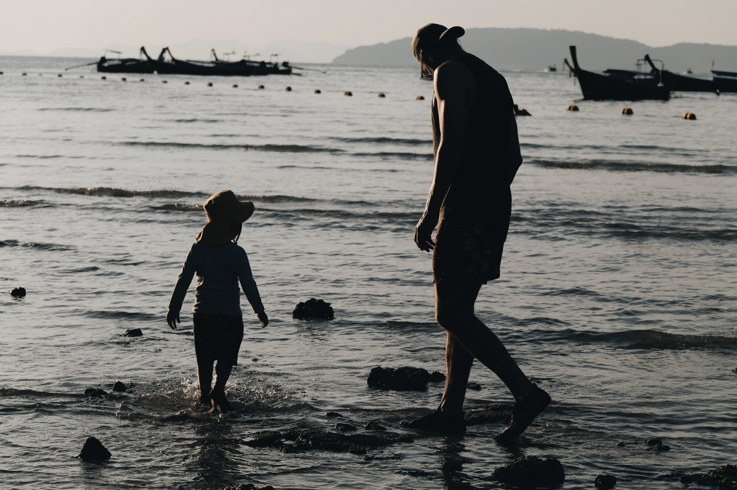 A silhouette of a man and a young girl walking along the shoreline at sunset or sunrise, with boats floating in the water in the background. Railey Beach Thailand