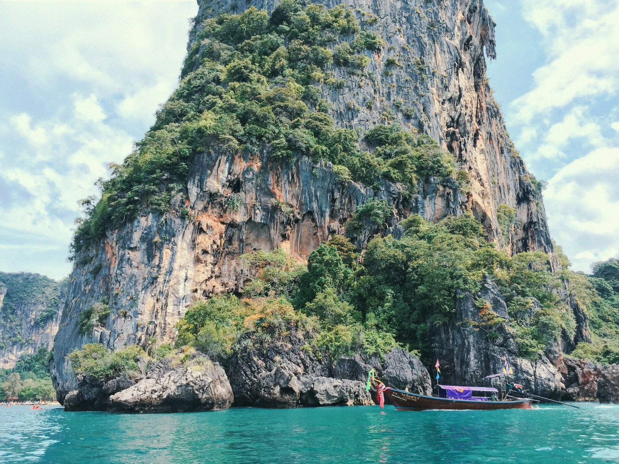 A boat floating near large rocky cliffs covered with green vegetation in a turquoise body of water under a cloudy sky.