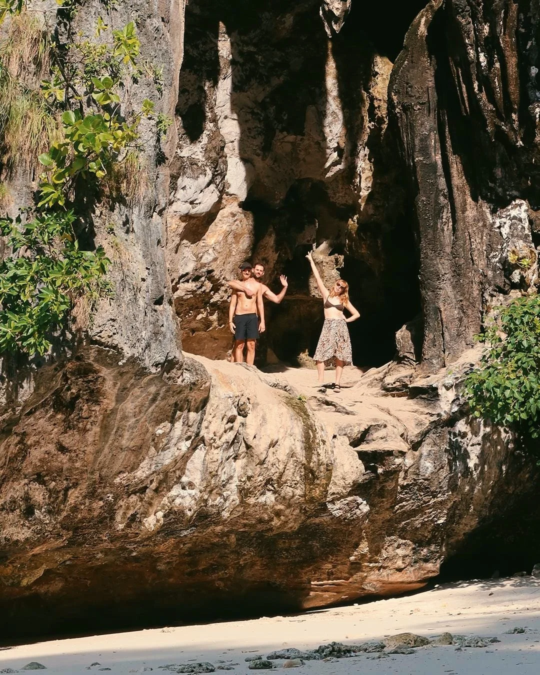 Three people standing on a large rock formation near a cave at the beach, with one man shirtless and one woman in a floral skirt and top waving, surrounded by some green foliage.
