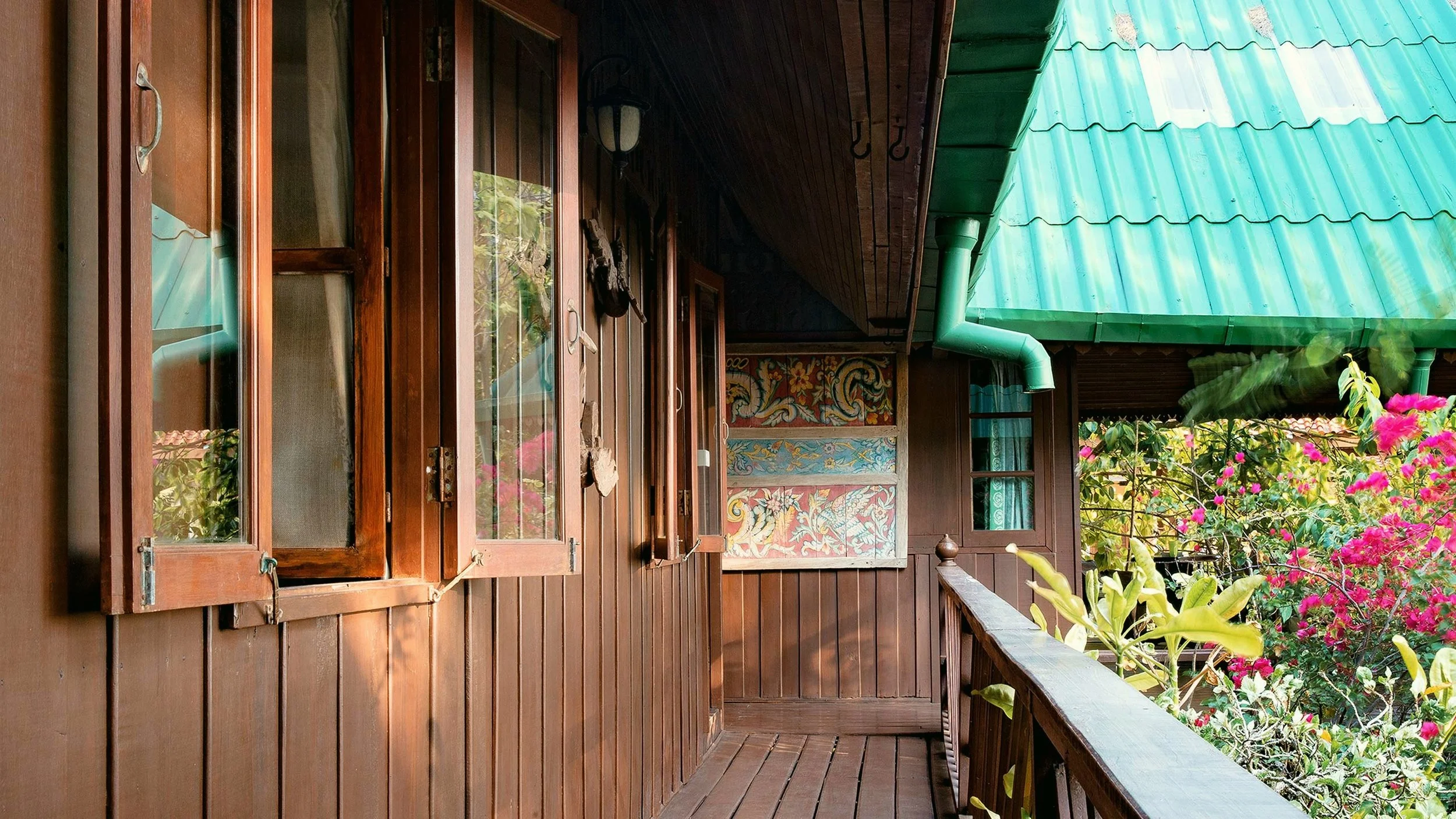 Wooden balcony of a house with open windows, colorful wall art, green roof, and vibrant pink flowers in the garden.