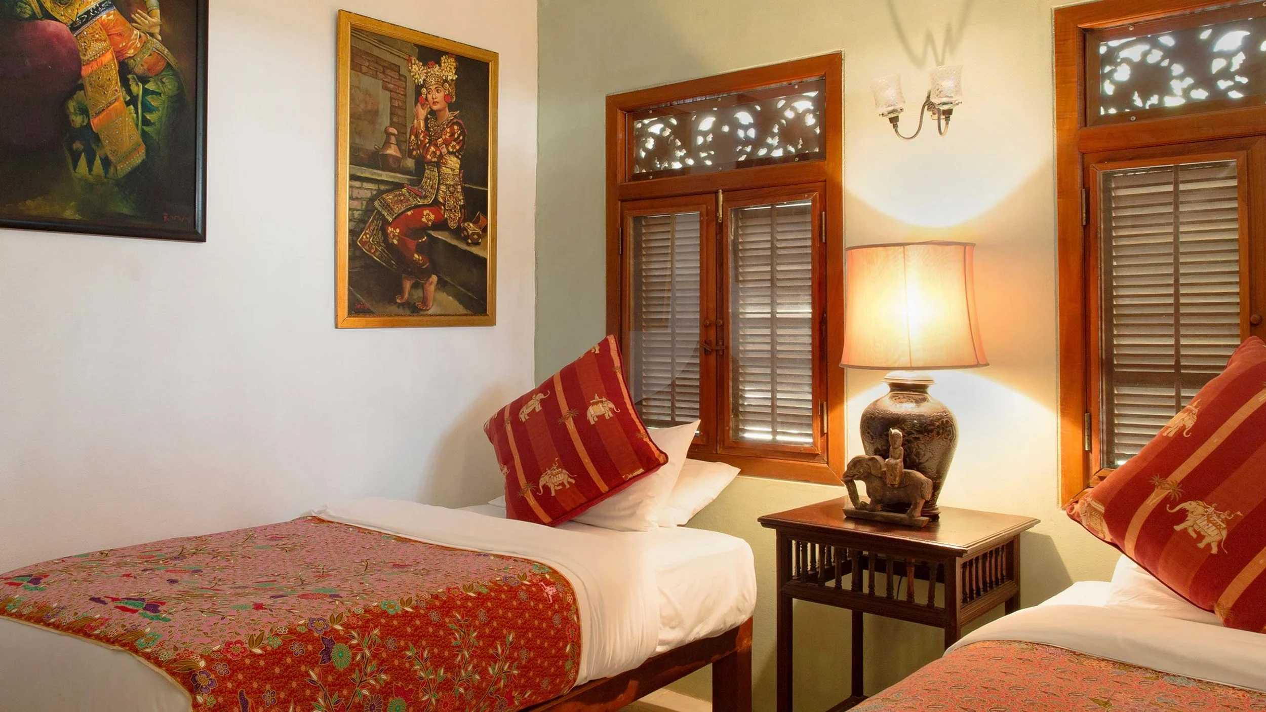 A cozy bedroom with two beds, colorful bedspreads, and matching pillows, illuminated by a table lamp on a wooden side table, with two wooden-framed windows and two paintings on the wall depicting traditional Thai art and culture.