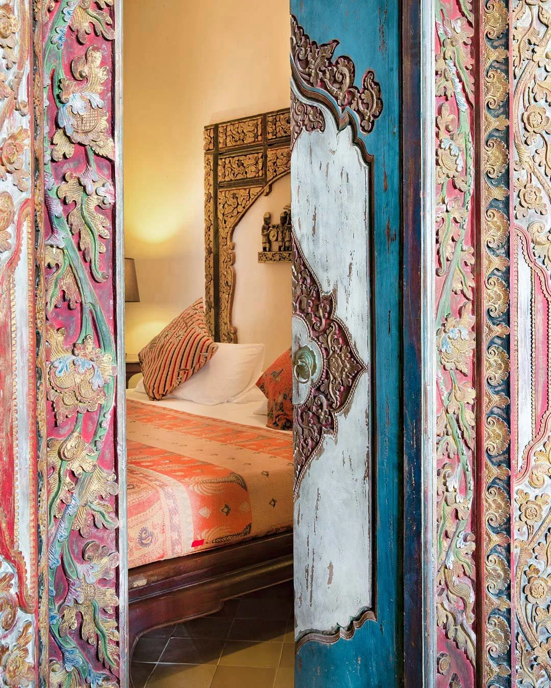 A colorful, ornately carved wooden door partially opens to reveal a bedroom with a bed, pillows, a nightstand with a lamp, and warm lighting, featuring traditional decorative elements and intricate detailing.