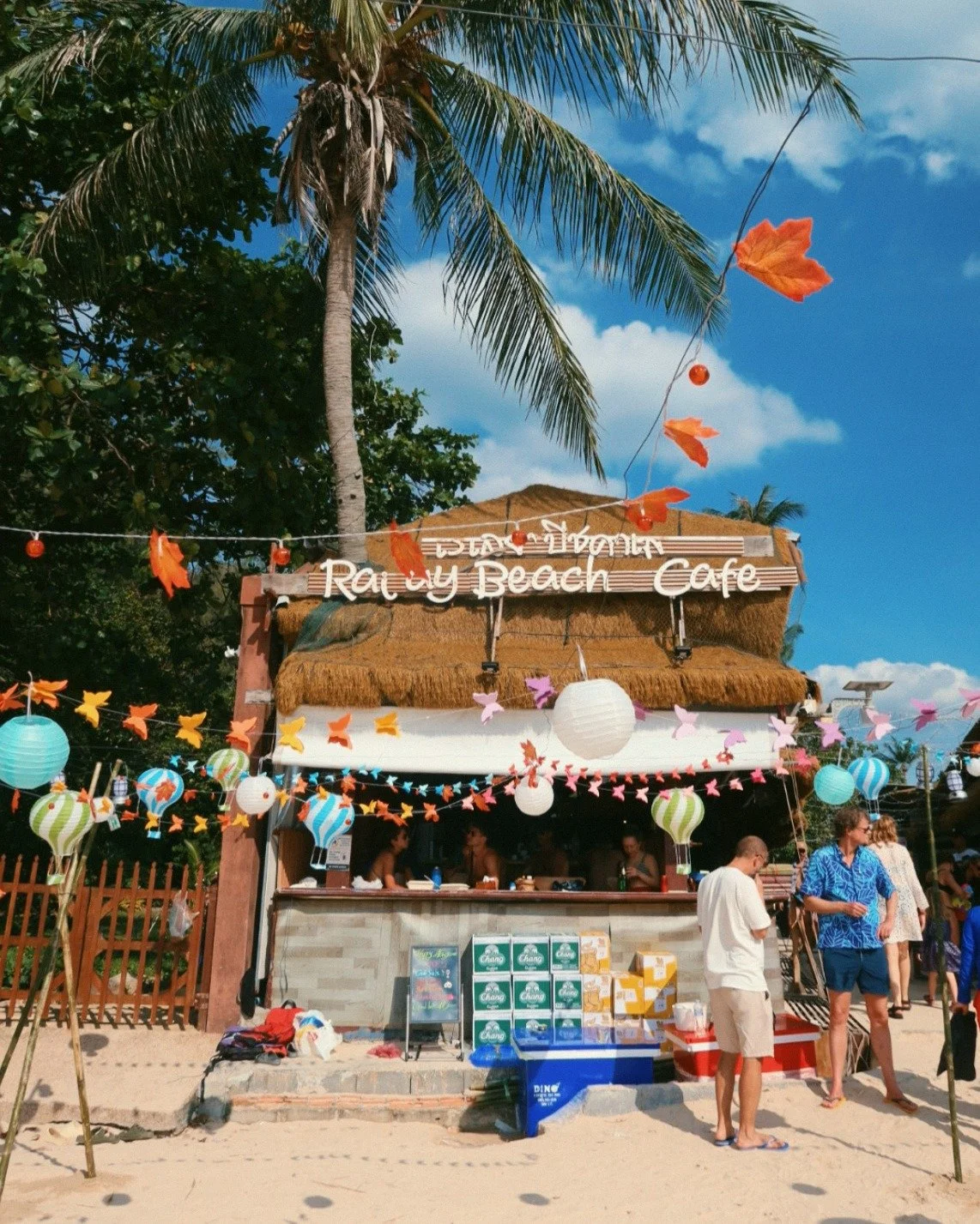Beachside cafe with colorful lanterns and string lights, palm trees, and people ordering and socializing under a thatched roof at Rayy Beach Cafe on a sunny day.