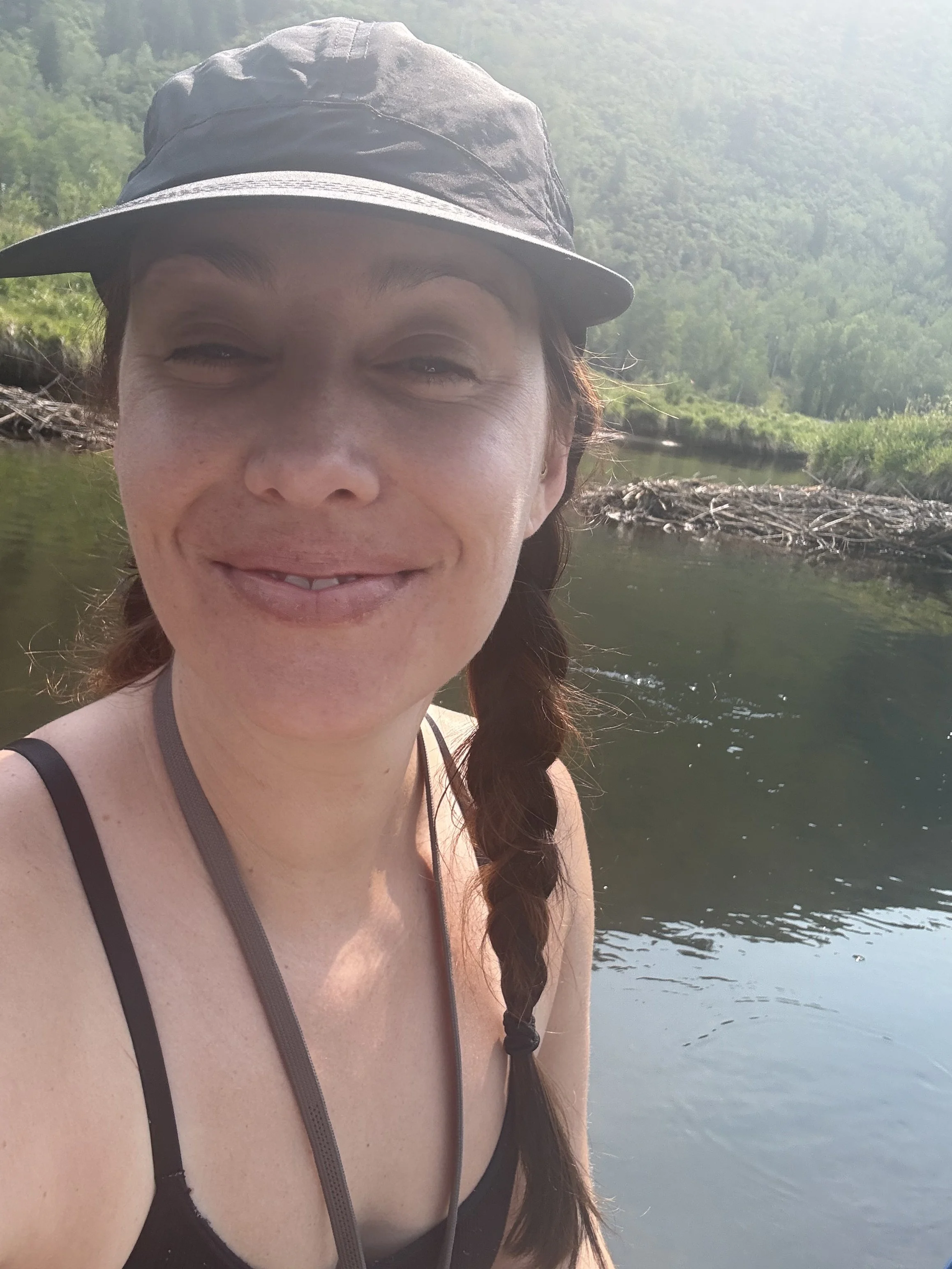 A woman with long braided hair, wearing a black cap and a black tank top, smiling outdoors near a river with green trees and hills in the background.
