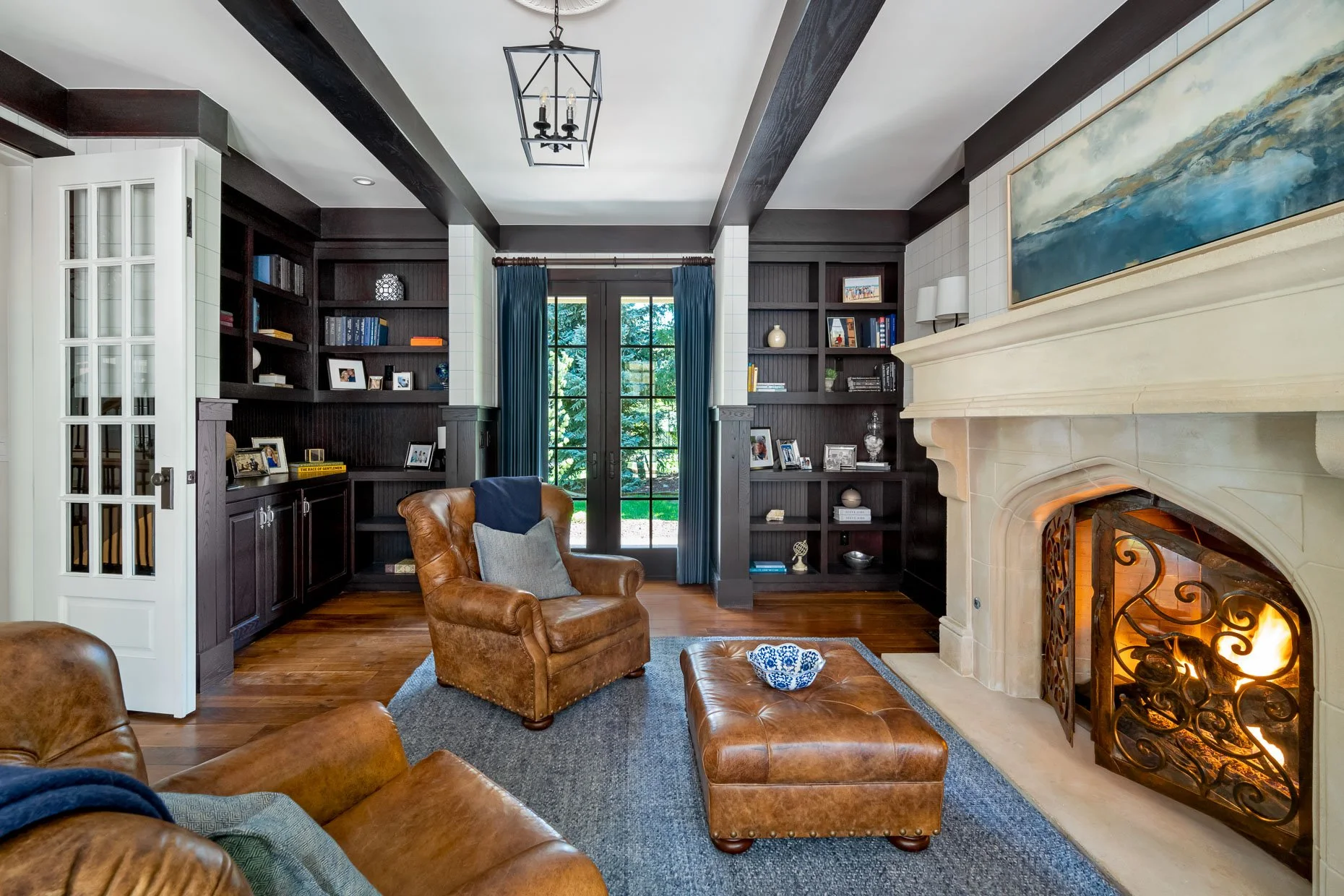 Study with a stone fireplace, leather armchair and ottoman, built-in dark wood bookshelves, blue curtains, and a door leading outside with trees visible.