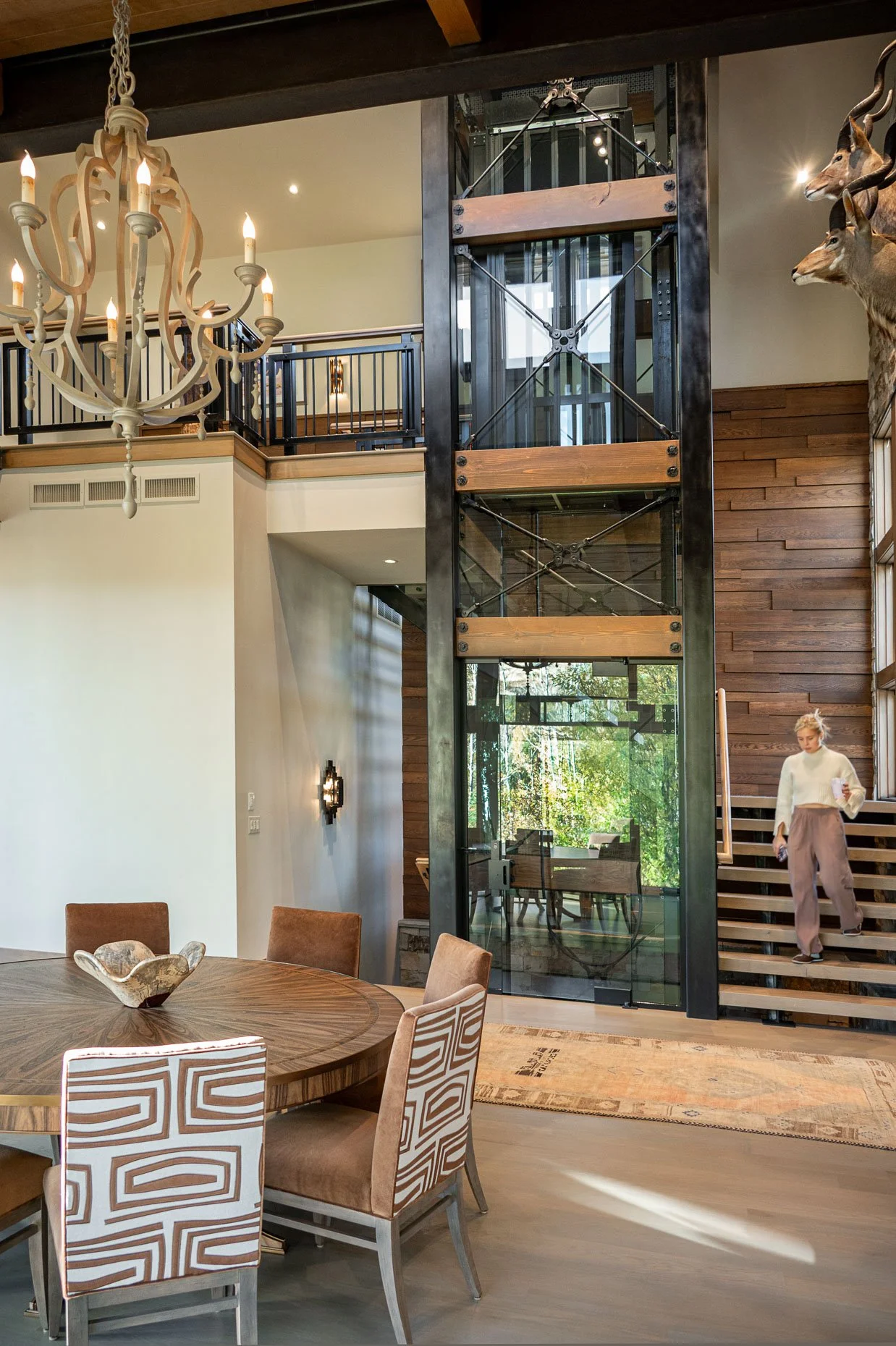 Denver interior design photography – Interior of a modern home with a wooden dining table and chairs, a chandelier, a glass elevator, wooden wall panels, and a staircase with a woman descending, with deer head mounts on the wall.