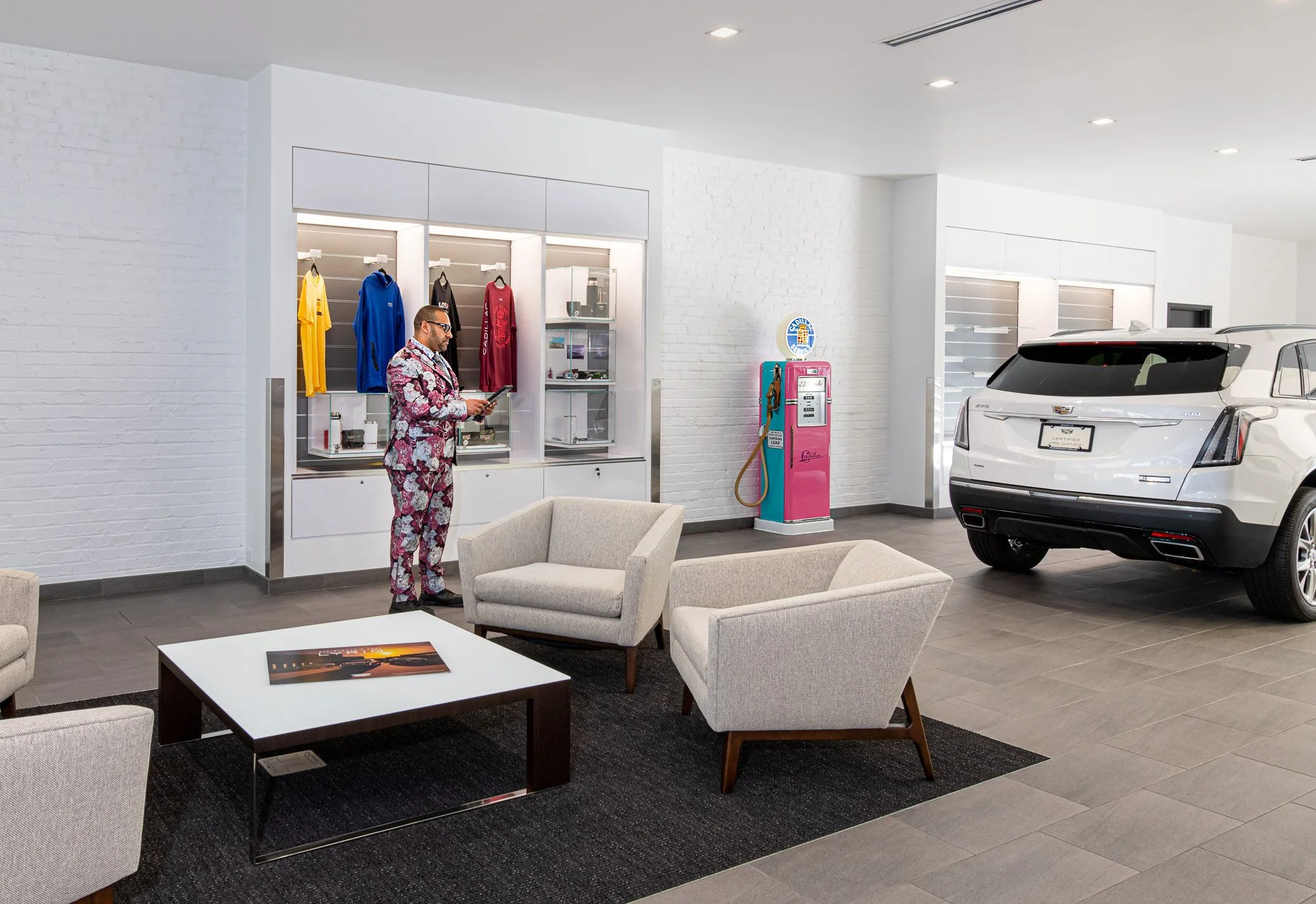 A modern car dealership showroom with a white SUV, a seating area with beige chairs and a white coffee table, a man in a floral suit looking at his phone, a vending machine in the background, and a display wall with colored sports jerseys.