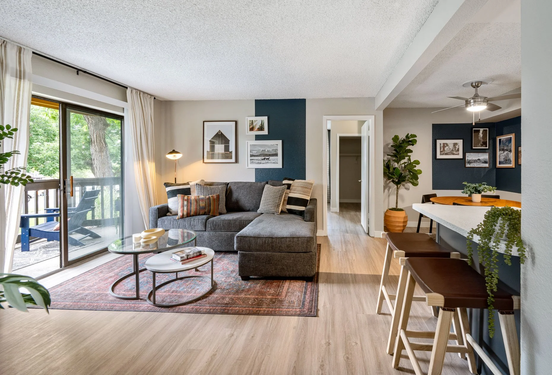 Living room with gray sectional sofa, glass coffee table, and sliding glass door leading to balcony with outdoor chairs, hardwood floor, and wall art on white wall with blue accent. Adjacent dining area with blue wall, round table, chairs, and indoor plant.