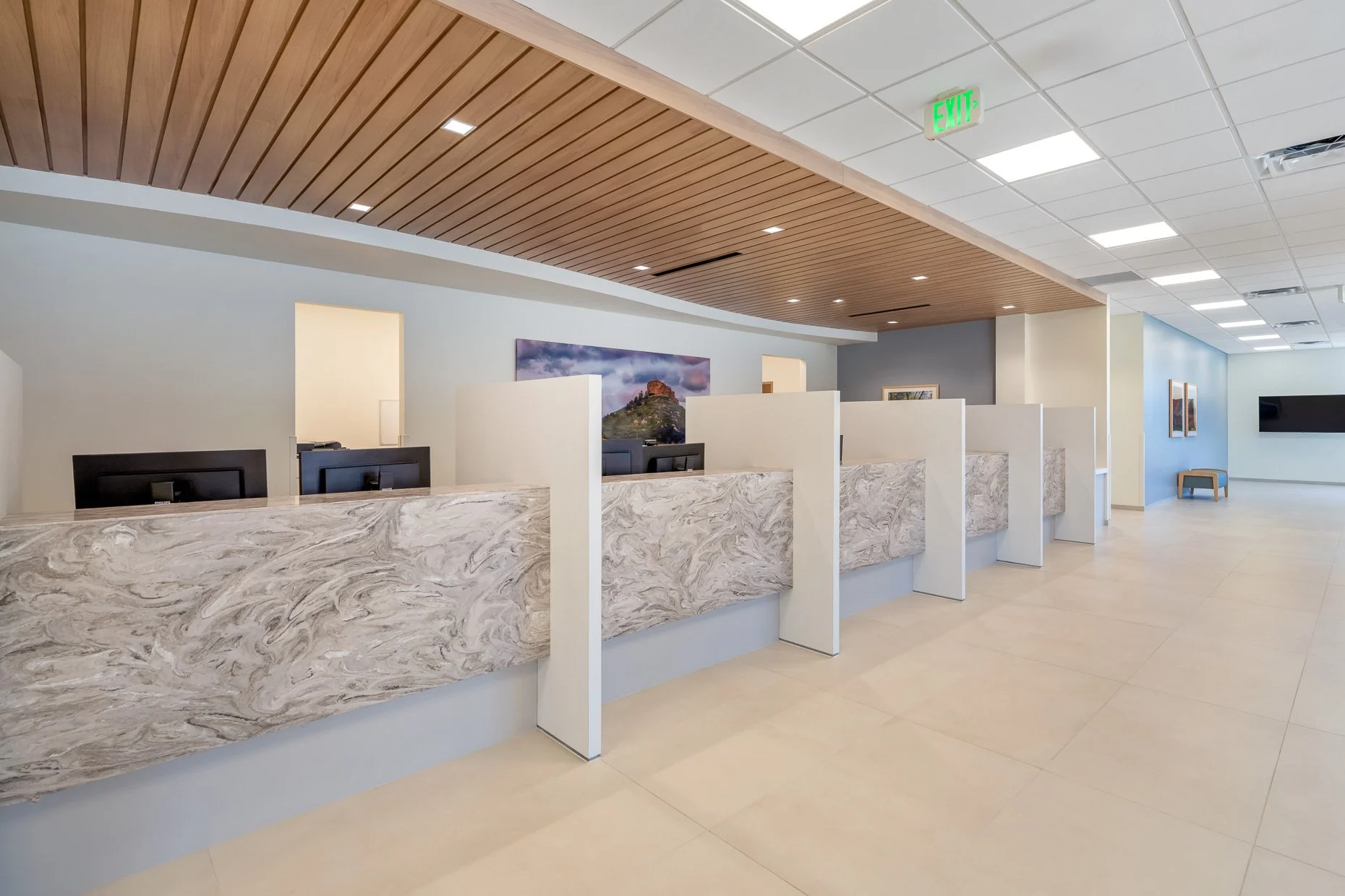 Empty office reception area with marble desks, computer monitors, wooden ceiling, framed artwork, and an emergency exit sign.