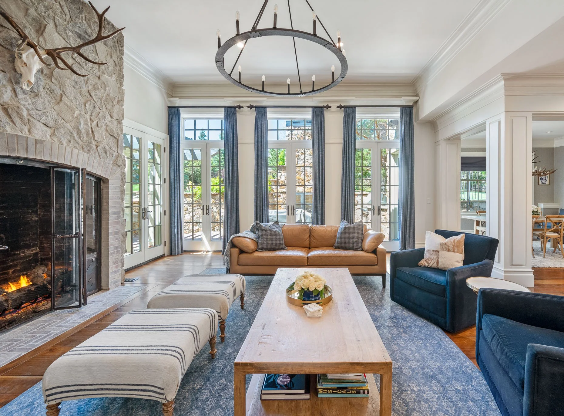 Living room with stone fireplace, tan leather sofa, navy armchairs, blue curtains, large windows, and a wooden coffee table with flowers, with a chandelier overhead.