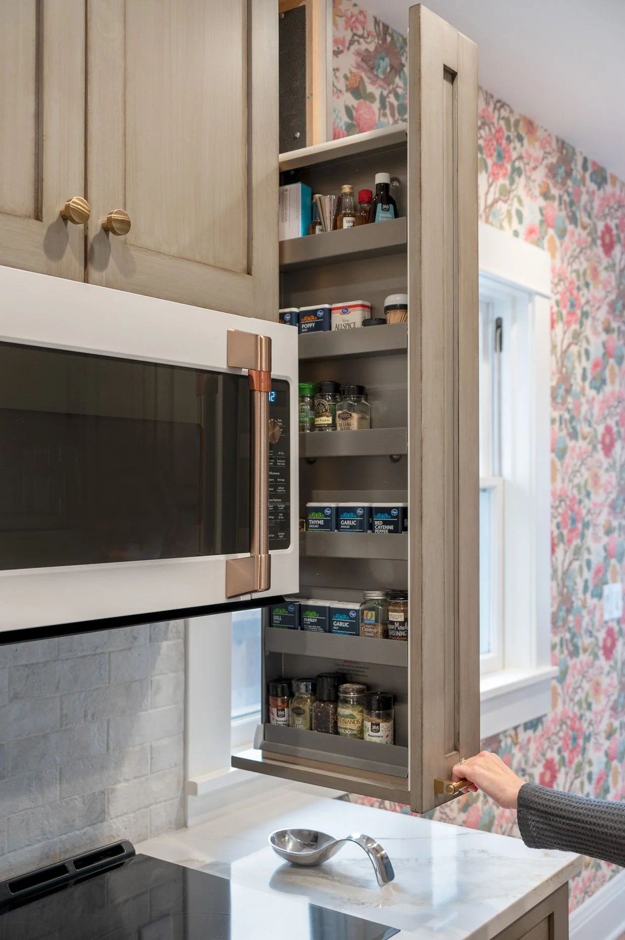 Person opening a kitchen cabinet filled with spices and condiments, with a microwave below and a marble countertop in the foreground.
