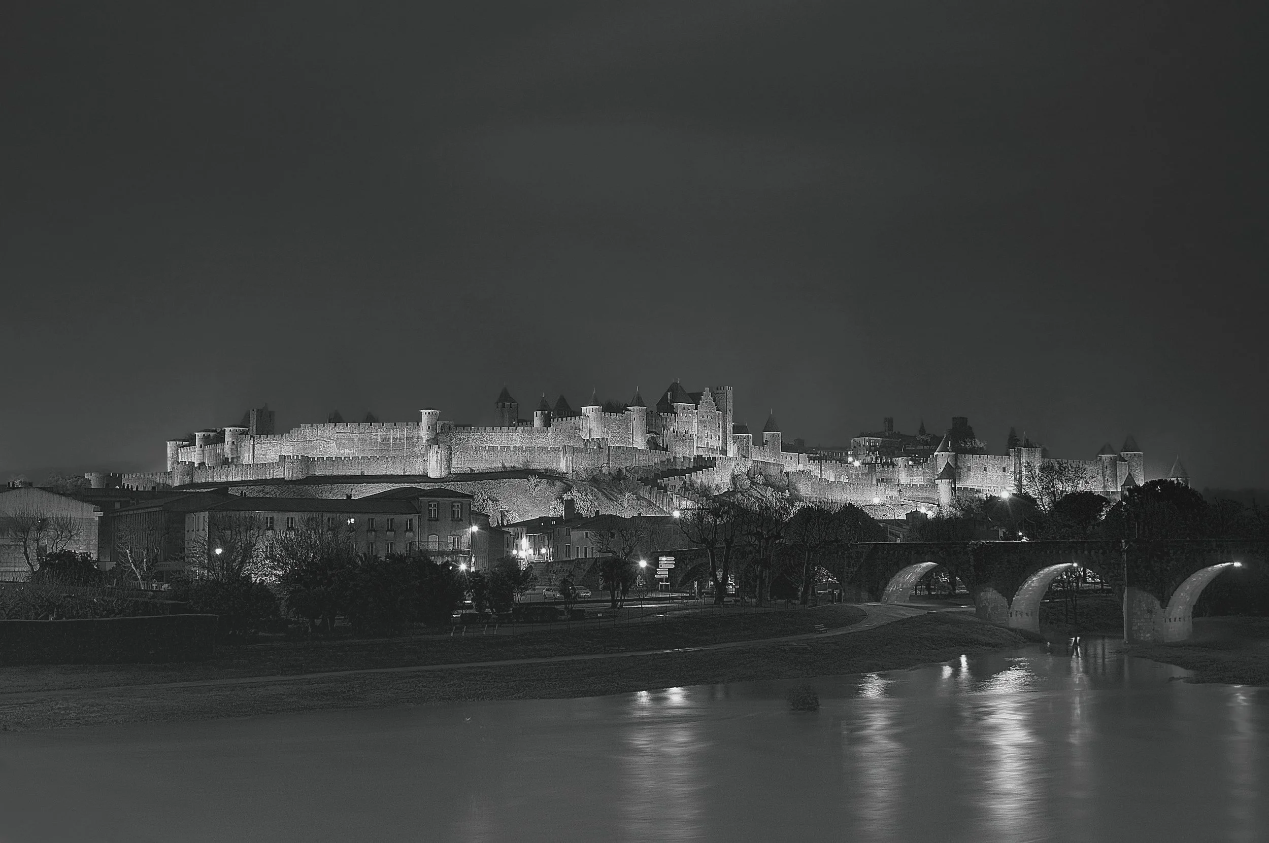 Une ville avec un château fortifié en arrière-plan, des bâtiments résidentiels et un pont au premier plan, toutes illuminées la nuit en noir et blanc.