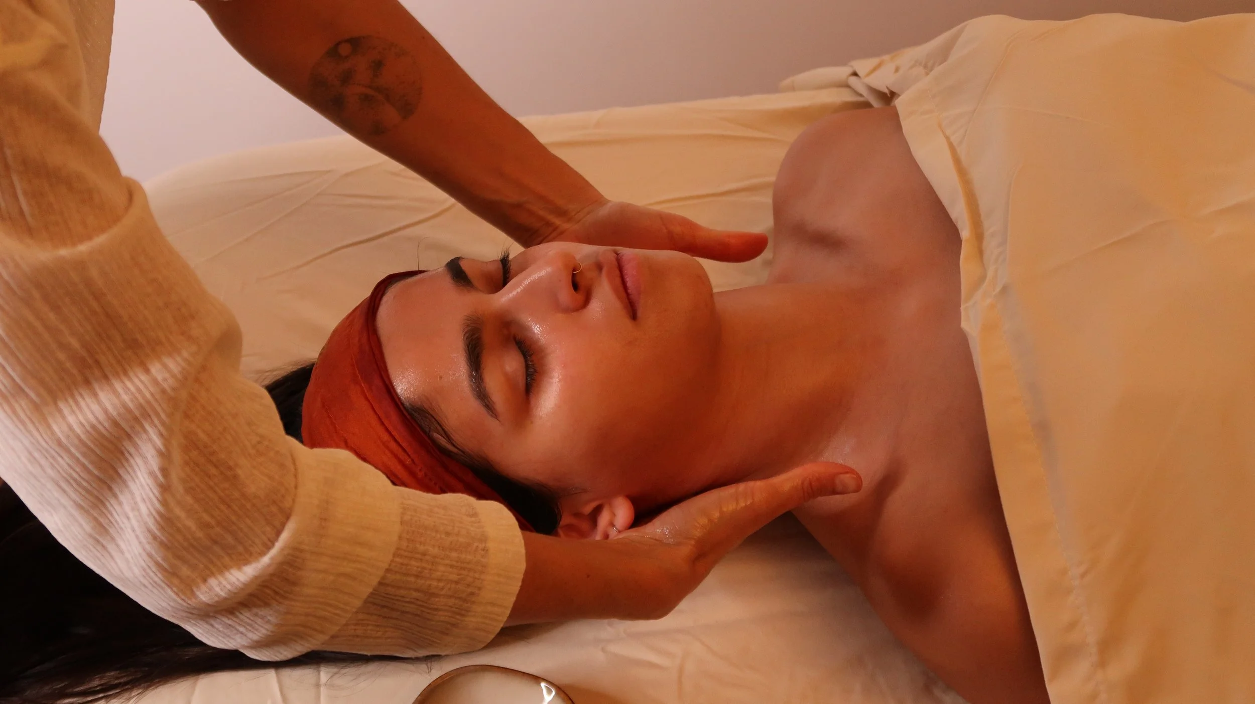 Femme recevant un massage du visage en position allongée, yeux fermés, avec un drap beige et une serviette sur la tête.