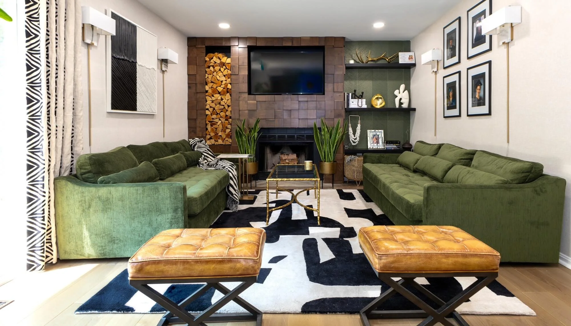 Living room with two green sofas, two brown leather ottomans, a black and white patterned rug, a wall-mounted TV, a fireplace with wood logs, plants, wall art, and decorative objects.