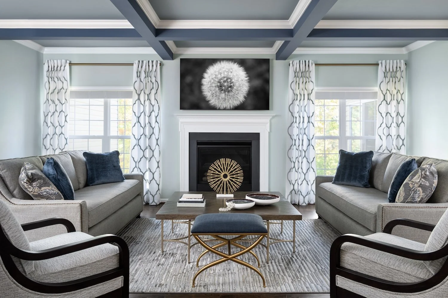 Living room with two gray sofas, two armchairs, a wooden coffee table, and a fireplace with a black frame. Decor includes pillows on the sofas, a modern abstract gold decorative piece on the table, and black-and-white artwork of a dandelion above the fireplace. Large windows with patterned curtains let in natural light.