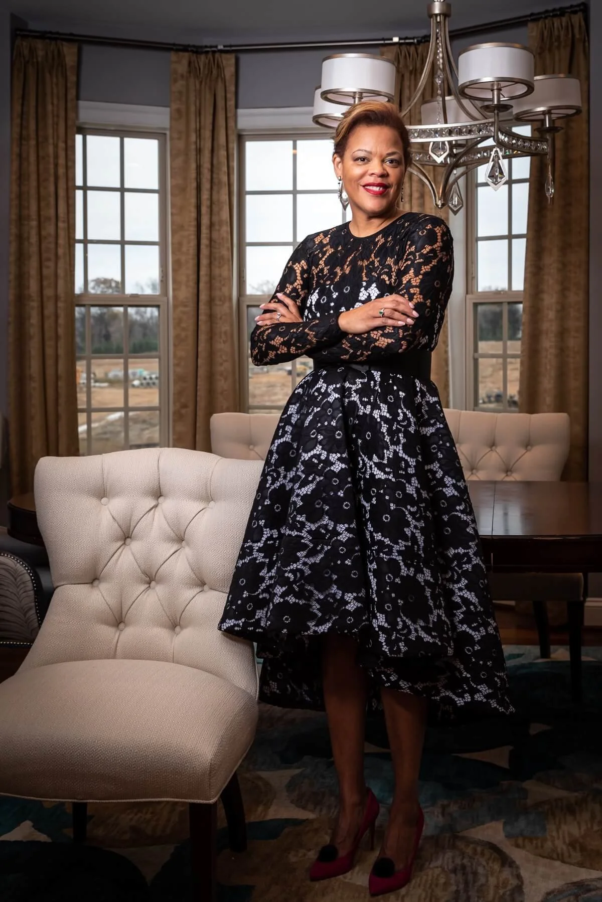 Monique stands confidently in a room with large windows and while dressed in a black lace dress with a floral pattern. 
